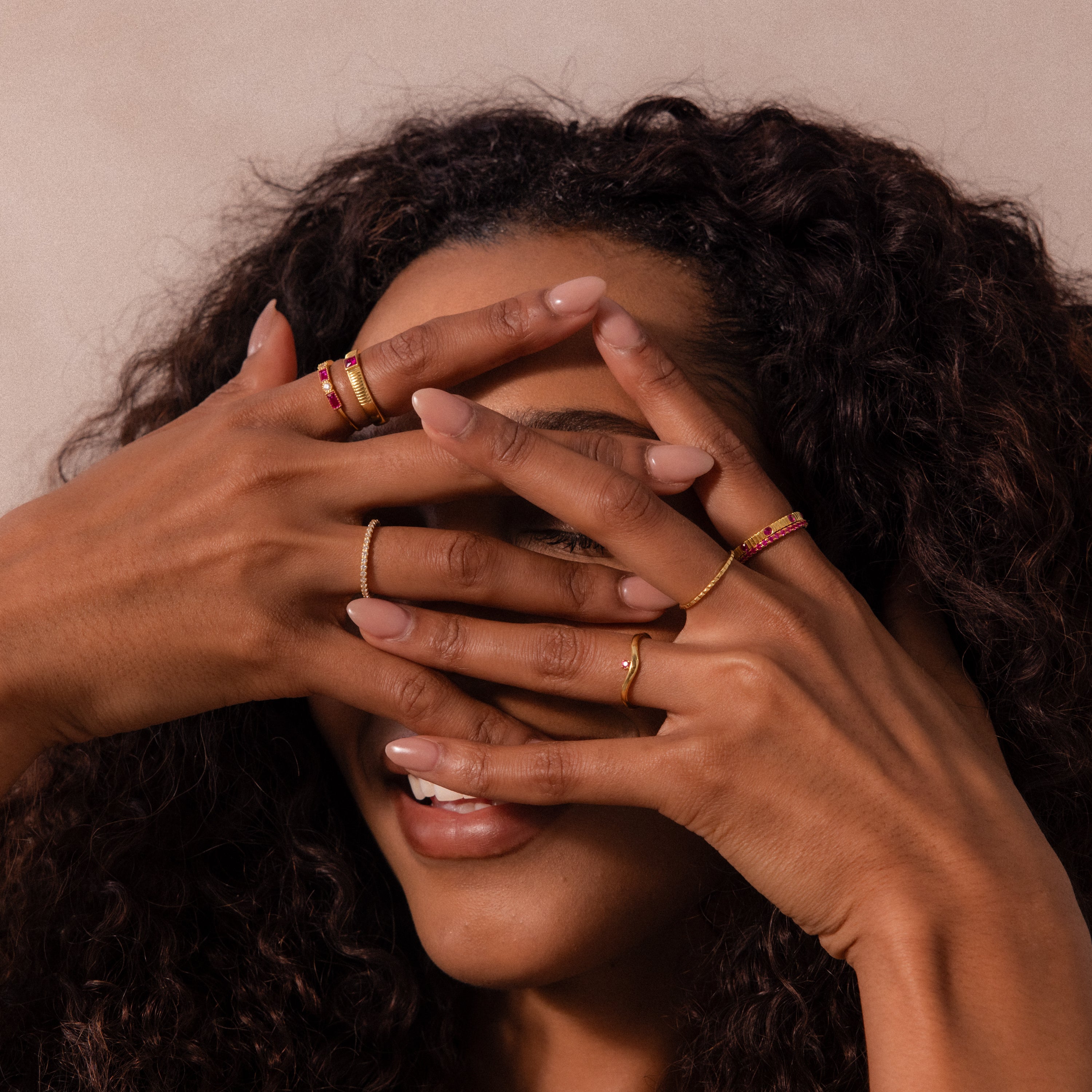 A smiling woman with curly hair covers her face with her hands, highlighting gold rings, including the Ribbed Garnet Band on her fingers.