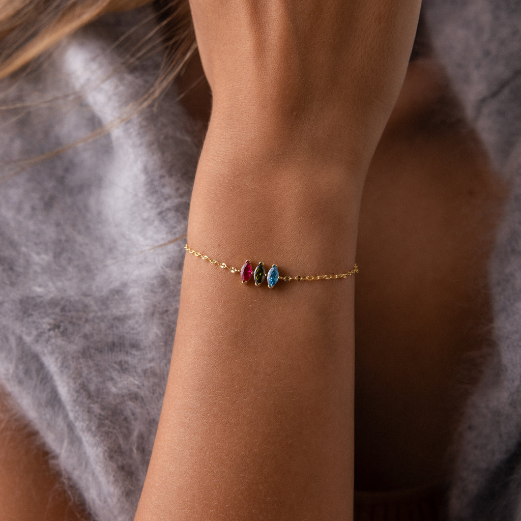 A Multiple Marquise Birthstone Bracelet featuring red, blue, and clear stones adorns a woman's wrist, beautifully matching her soft gray sweater—a stunning customizable birthstone jewelry piece.