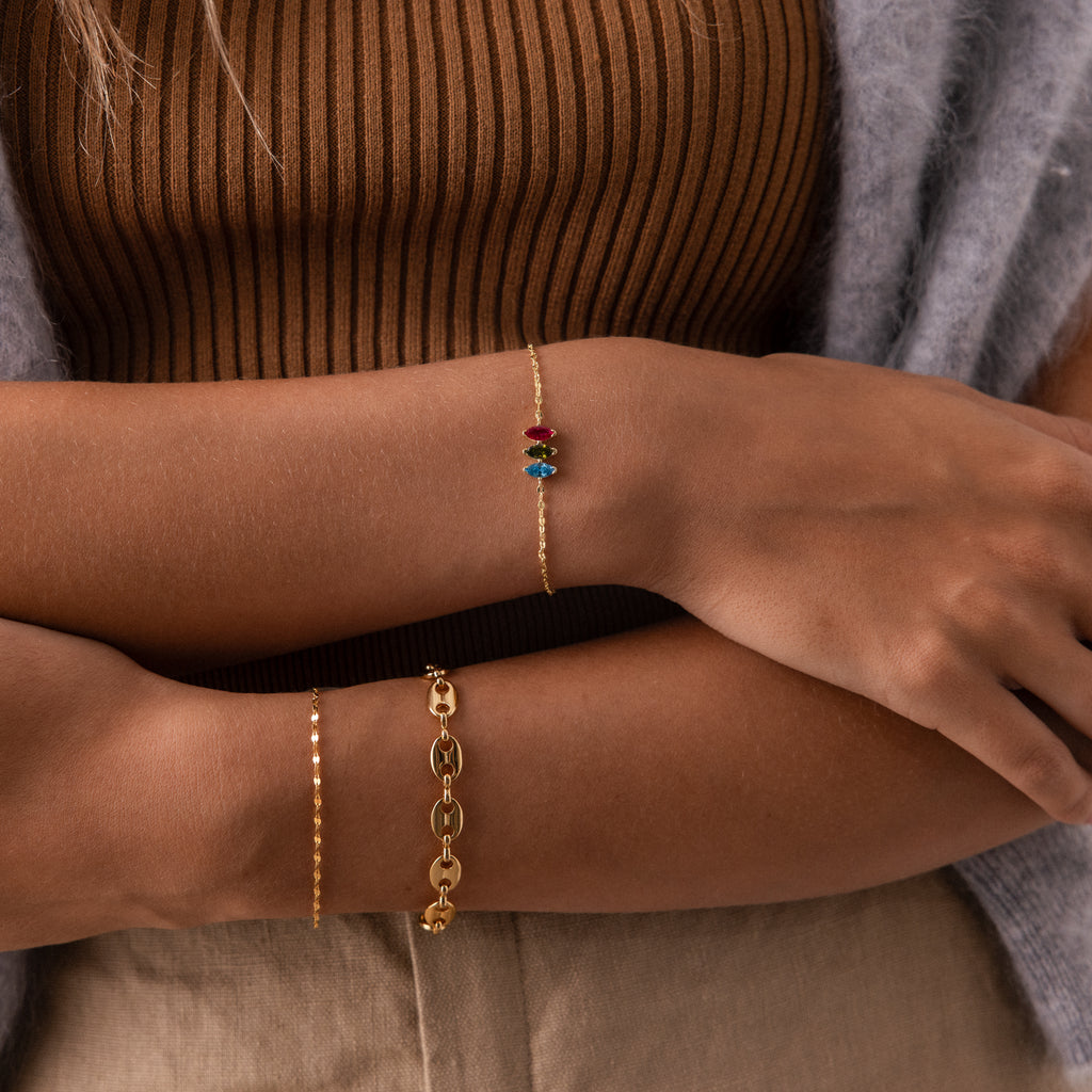 A person wearing a gold bracelet, the Multiple Marquise Birthstone Bracelet, and a beaded chain bracelet, dressed in a brown ribbed top and beige pants.