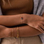 A person wearing a gold bracelet, the Multiple Marquise Birthstone Bracelet, and a beaded chain bracelet, dressed in a brown ribbed top and beige pants.