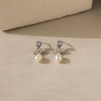 Aquamarine Pearl Drop Earrings in Silver, adorned with blue stones, clear crystals, and dangling pearls, exude vintage glamour on a beige surface with soft shadows.