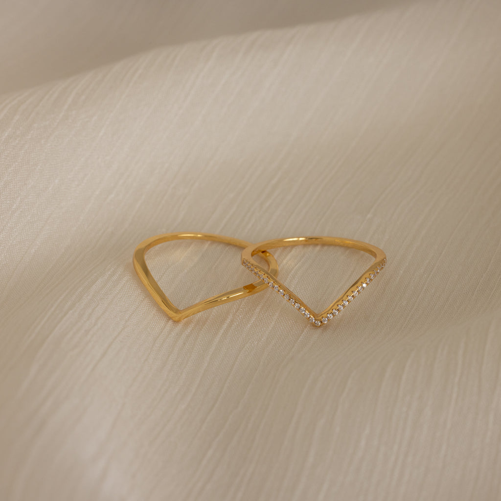 The Curve Stacking Set in 18K Gold features two elegant rings—one plain, one accented with small diamonds—styled together atop a soft beige fabric background.