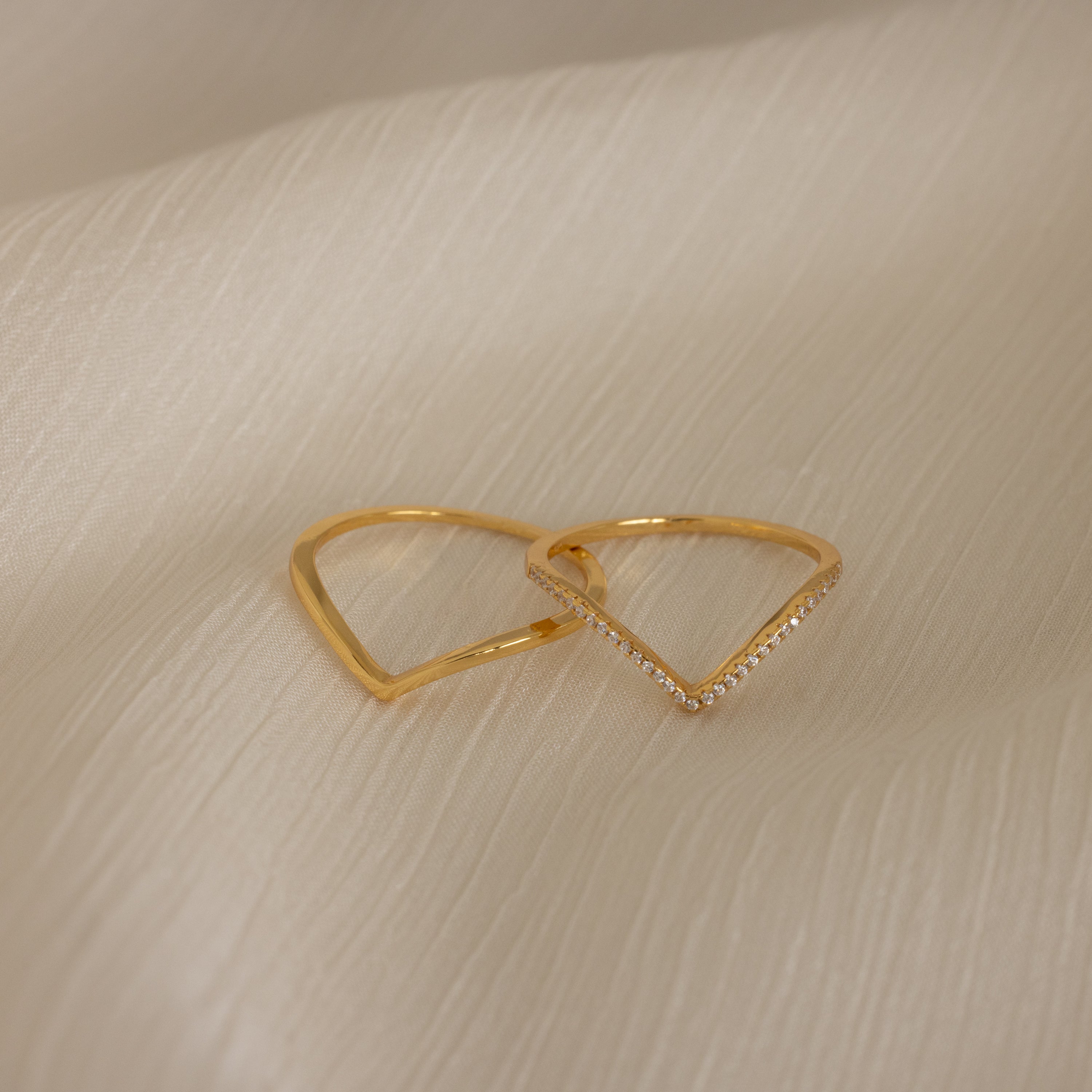 The Curve Stacking Set in 18K Gold features two elegant rings—one plain, one accented with small diamonds—styled together atop a soft beige fabric background.
