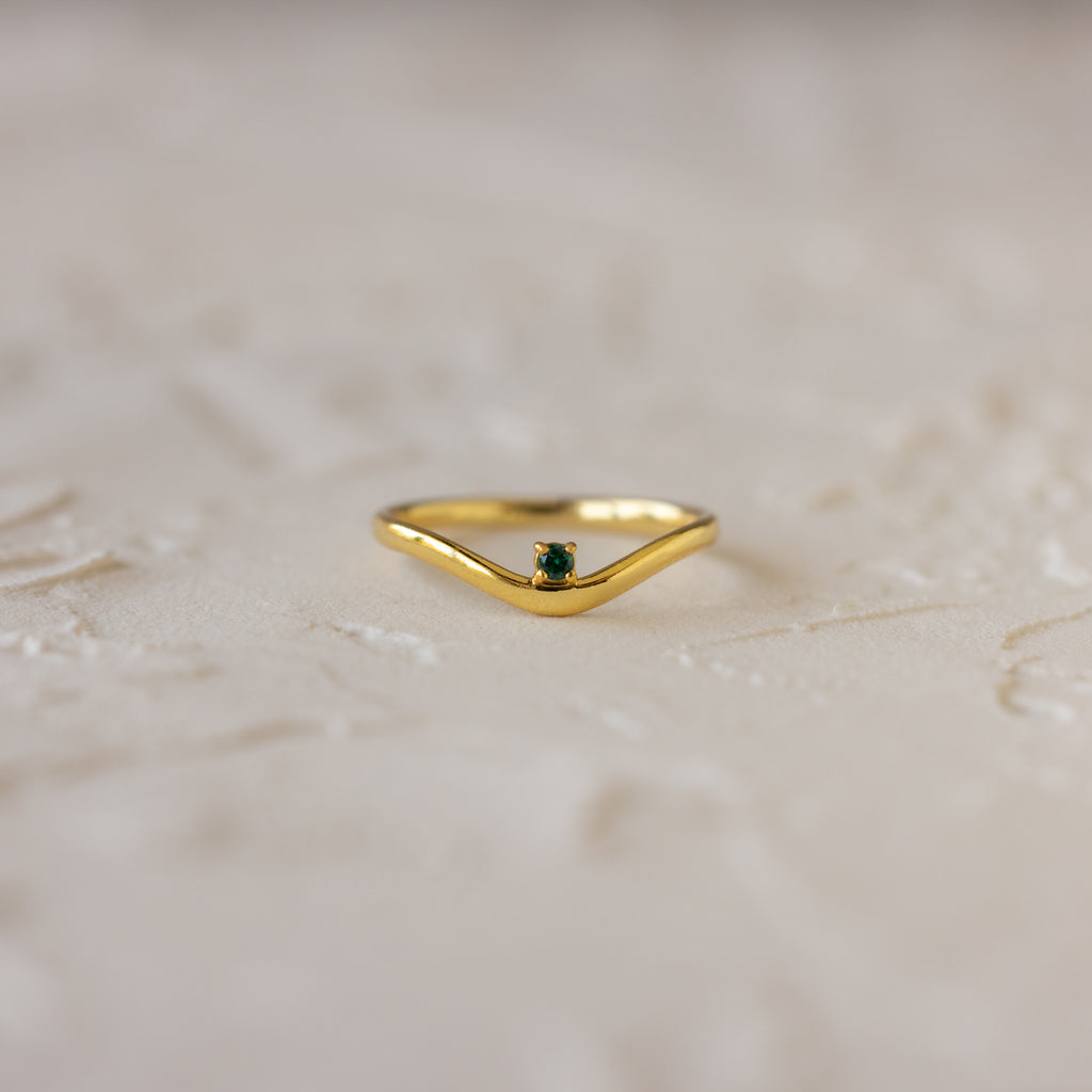 The Emerald Drop Curve Ring is a delicate gold stacking ring with a small green gemstone, perfect for wearing alone or stacked, shown on a textured light surface.
