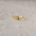 The Emerald Drop Curve Ring is a delicate gold stacking ring with a small green gemstone, perfect for wearing alone or stacked, shown on a textured light surface.
