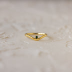 The Emerald Drop Curve Ring is a delicate gold stacking ring with a small green gemstone, perfect for wearing alone or stacked, shown on a textured light surface.