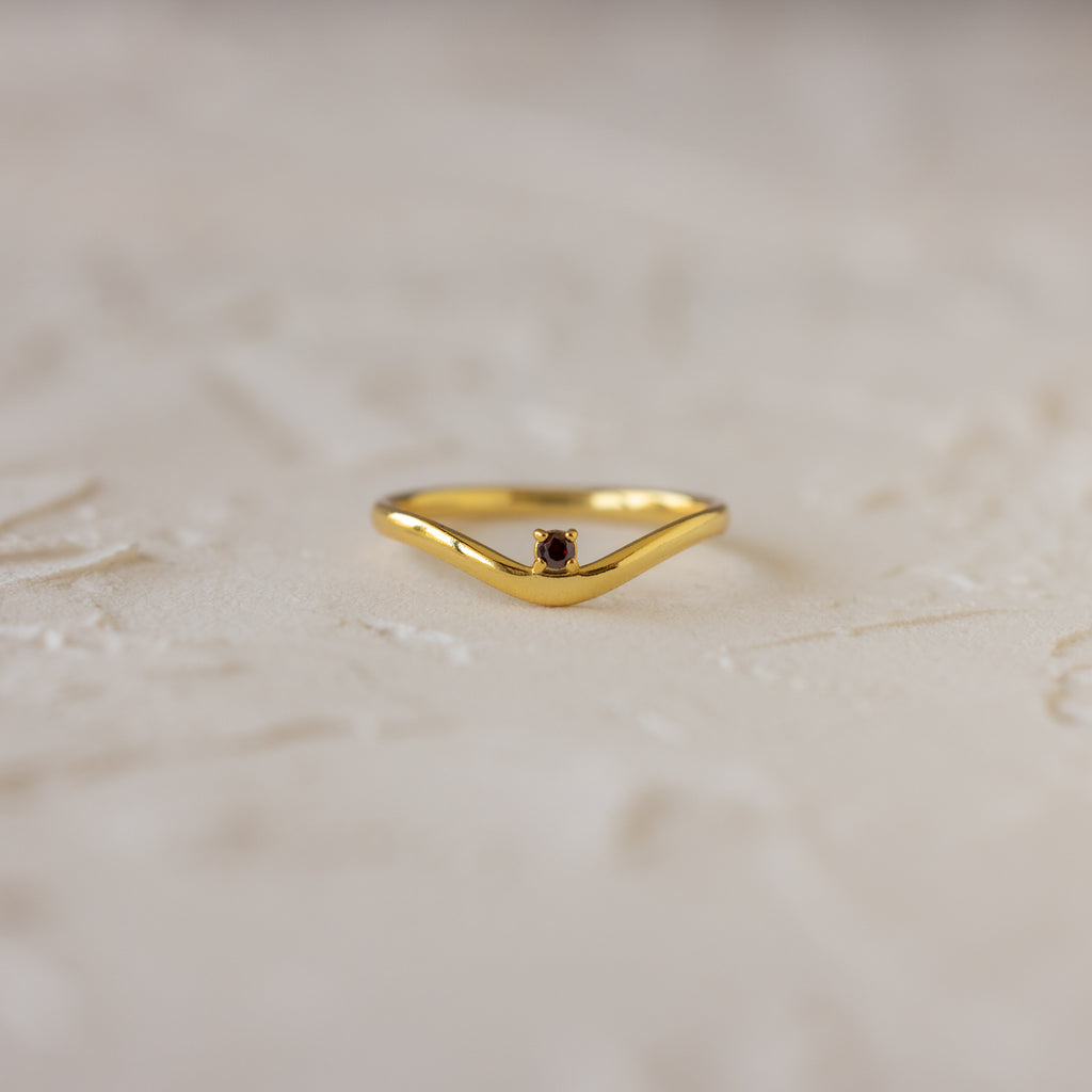 The Garnet Drop Curve Ring features a delicate gold curve and a small red garnet at its center, perfect for stacking, displayed on a light textured surface.