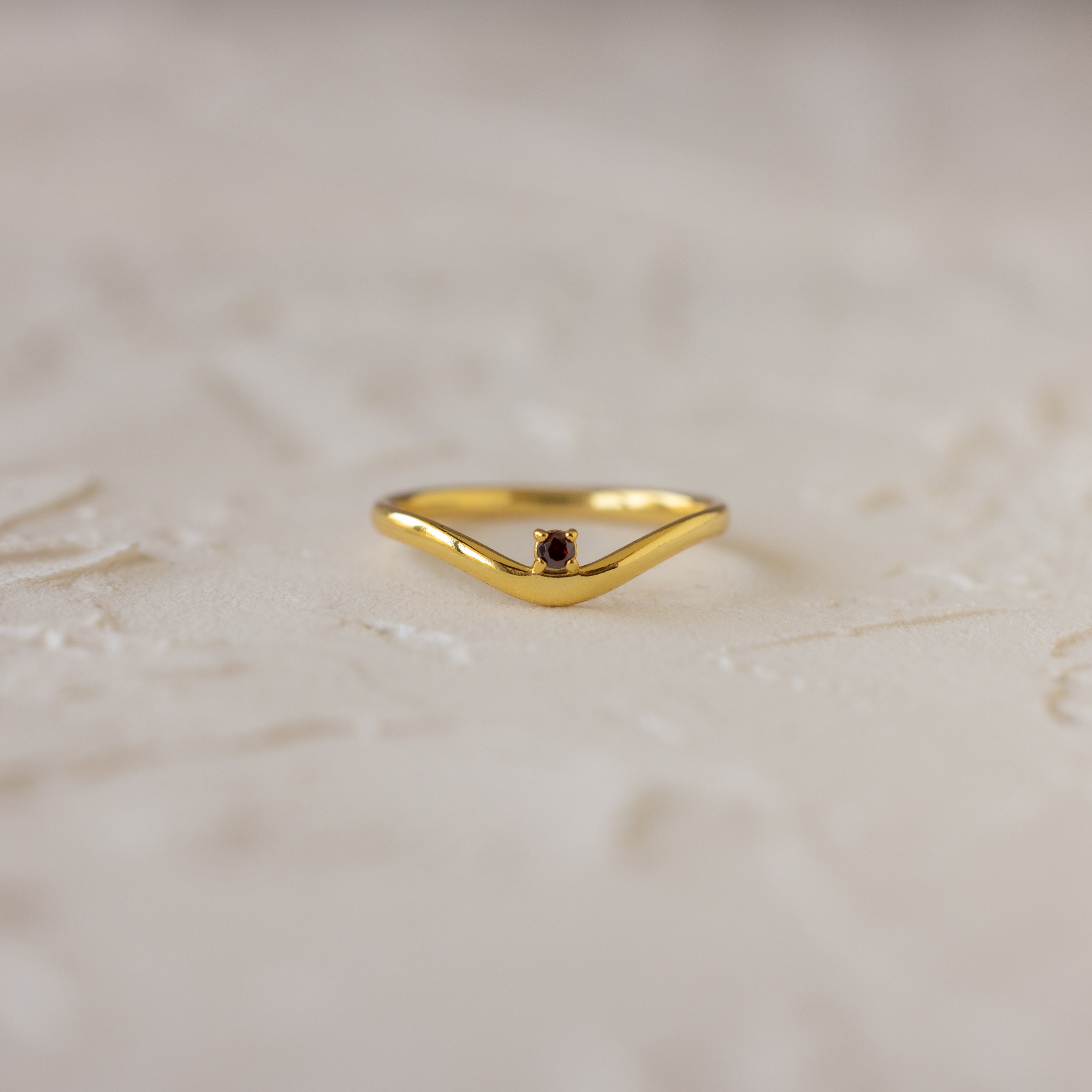 The Garnet Drop Curve Ring features a delicate gold curve and a small red garnet at its center, perfect for stacking, displayed on a light textured surface.