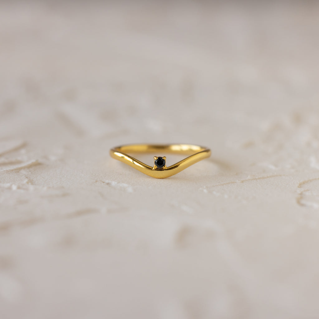The Onyx Drop Curve Ring is a delicate gold stacking ring with a small dark onyx gemstone, perfect as a subtle birthstone accent and beautifully displayed on a textured light surface.