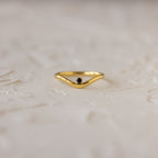 The Onyx Drop Curve Ring is a delicate gold stacking ring with a small dark onyx gemstone, perfect as a subtle birthstone accent and beautifully displayed on a textured light surface.