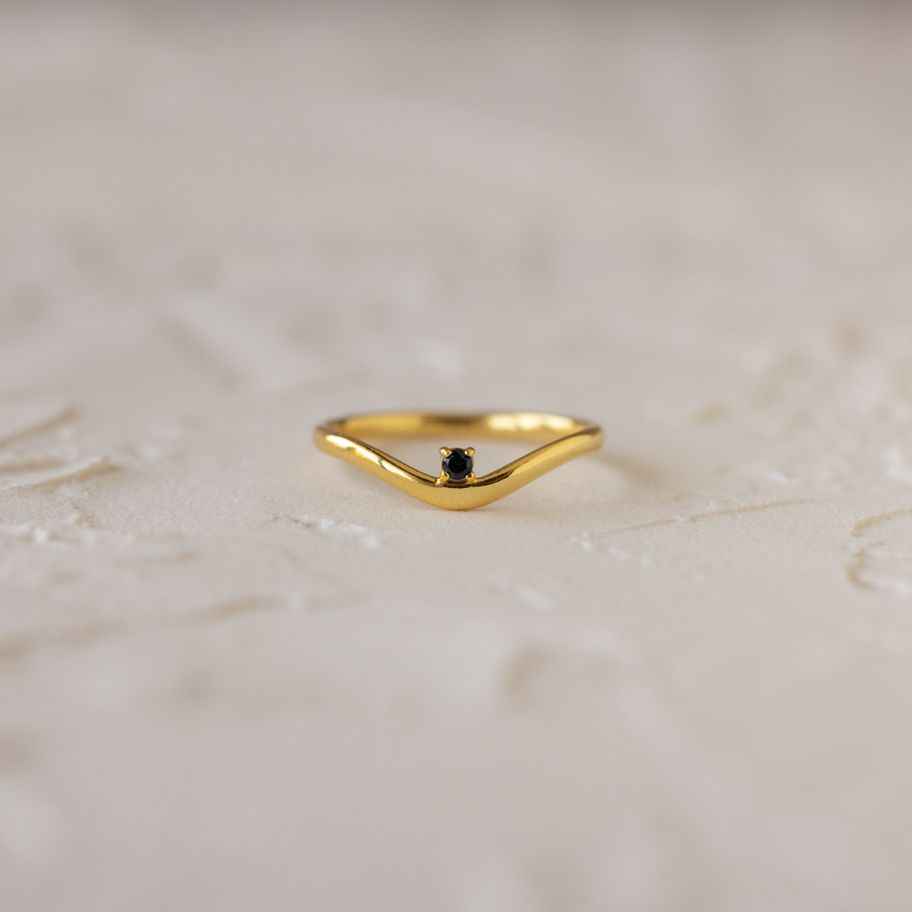 The Onyx Drop Curve Ring is a delicate gold stacking ring with a small dark onyx gemstone, perfect as a subtle birthstone accent and beautifully displayed on a textured light surface.