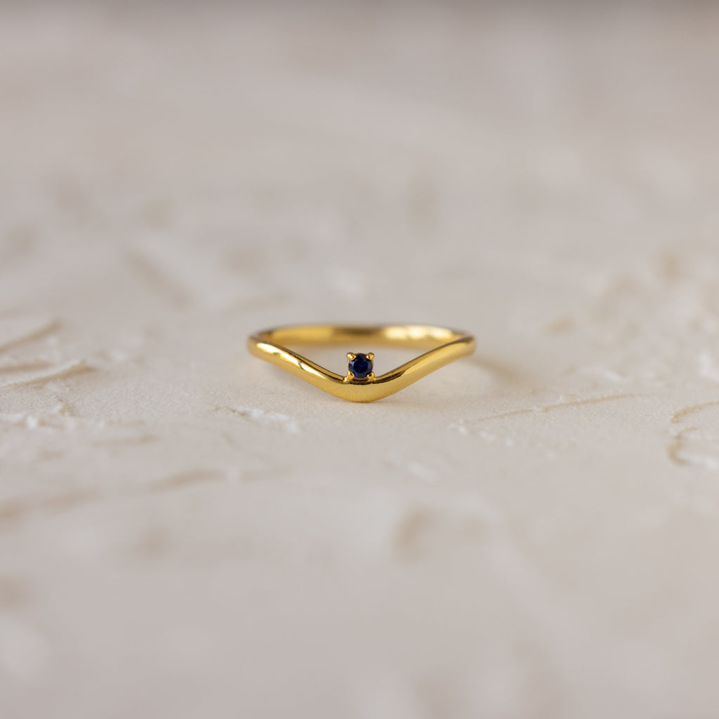 The Sapphire Drop Curve Ring, a gold stacking ring with a small dark sapphire gemstone, rests on a textured, light-colored surface.
