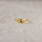 The Sapphire Drop Curve Ring, a gold stacking ring with a small dark sapphire gemstone, rests on a textured, light-colored surface.