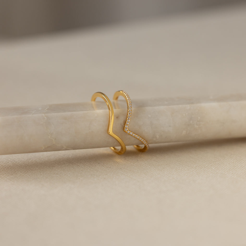 The Curve Stacking Set in 18K Gold features two slim zigzag bands—one plain, one with sparkling stones—resting on beige marble for a chic, stackable look.