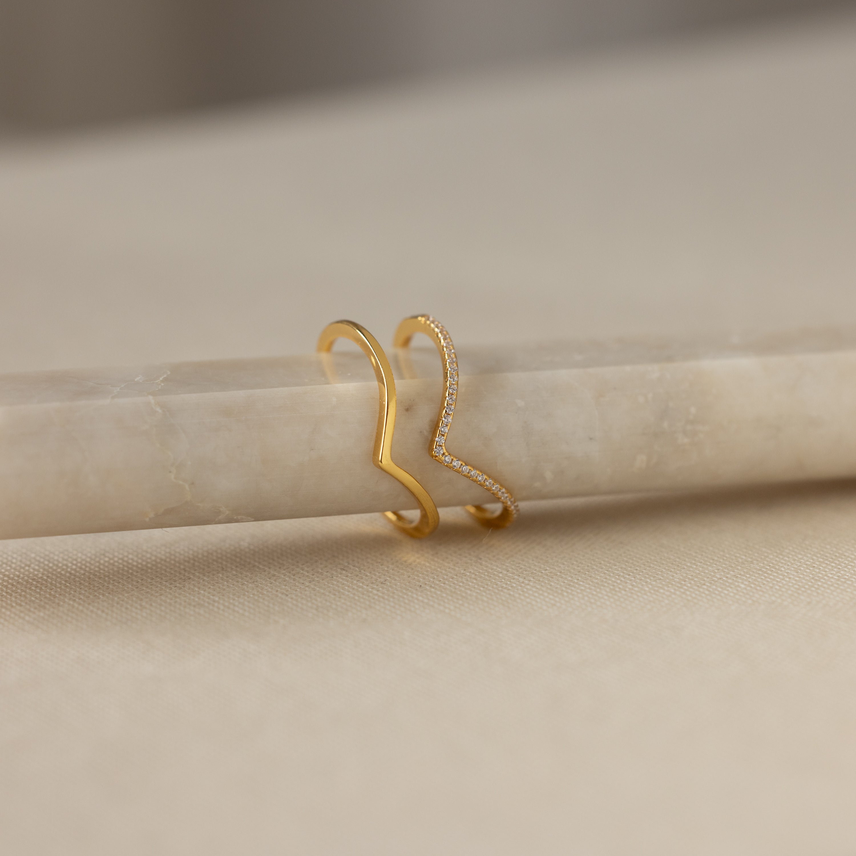 The Curve Stacking Set in 18K Gold features two slim zigzag bands—one plain, one with sparkling stones—resting on beige marble for a chic, stackable look.