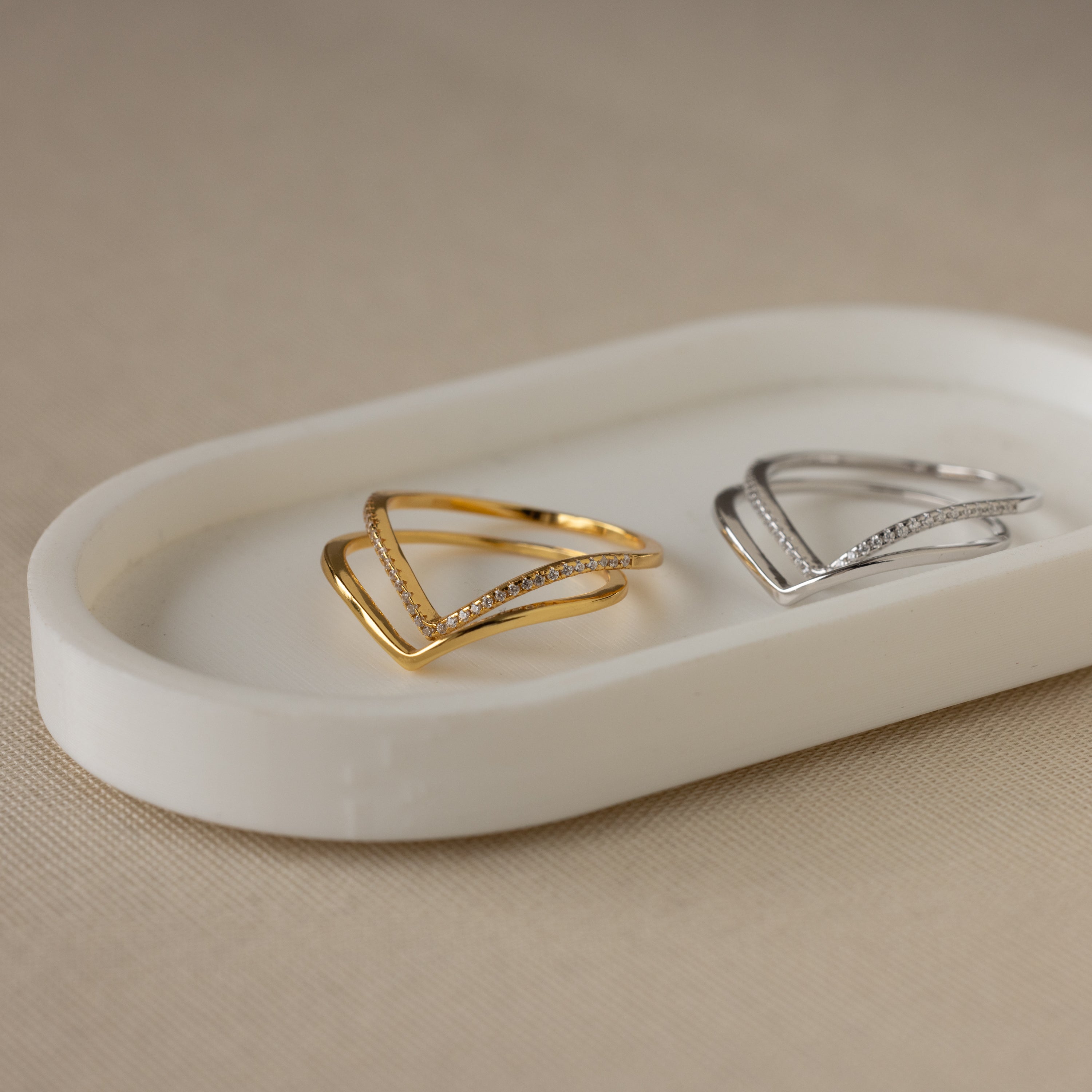 The Curve Stacking Set features two minimalist V-shaped thin band rings, one gold and one silver with small stones, displayed on a white tray.
