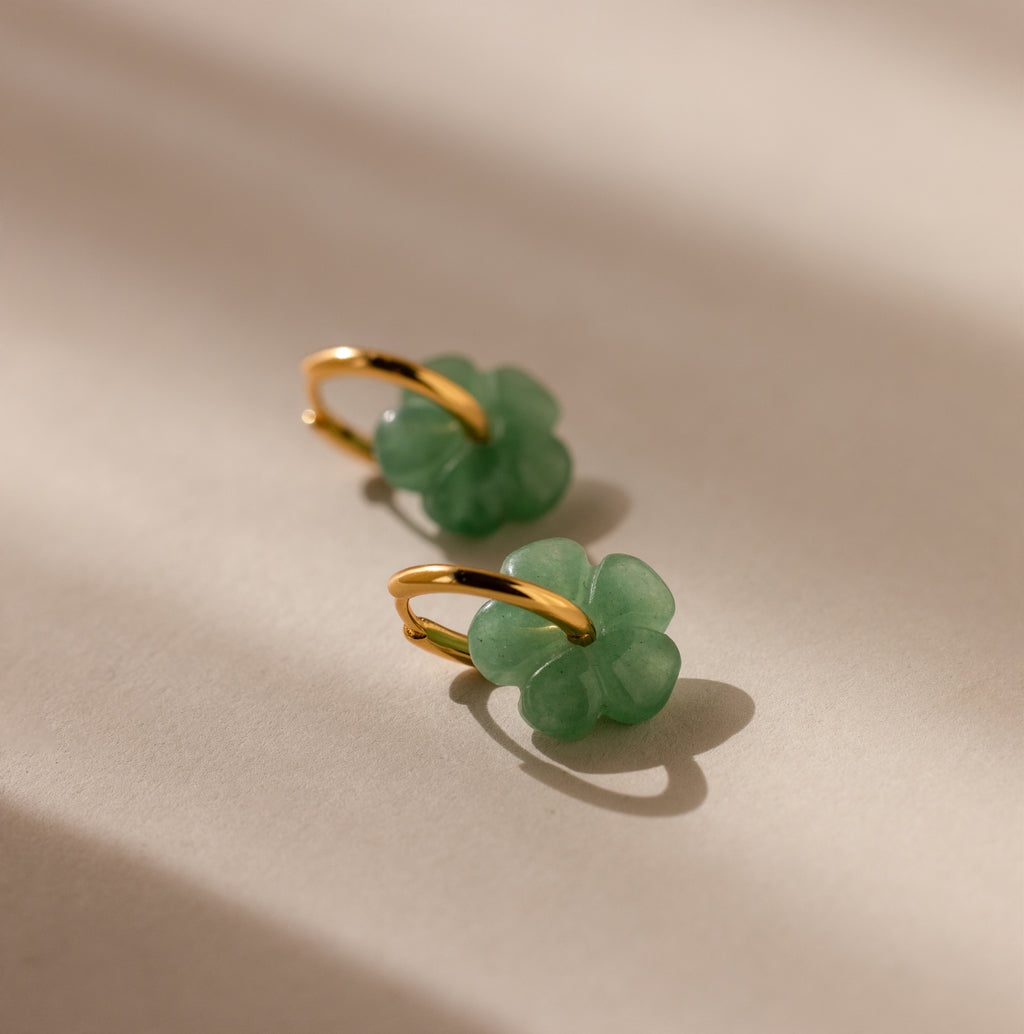 Jade Flower Hoops—gold hoop earrings with green jade flower accents—rest on a beige surface with soft shadows. Dainty and stylish, they make a meaningful gift.