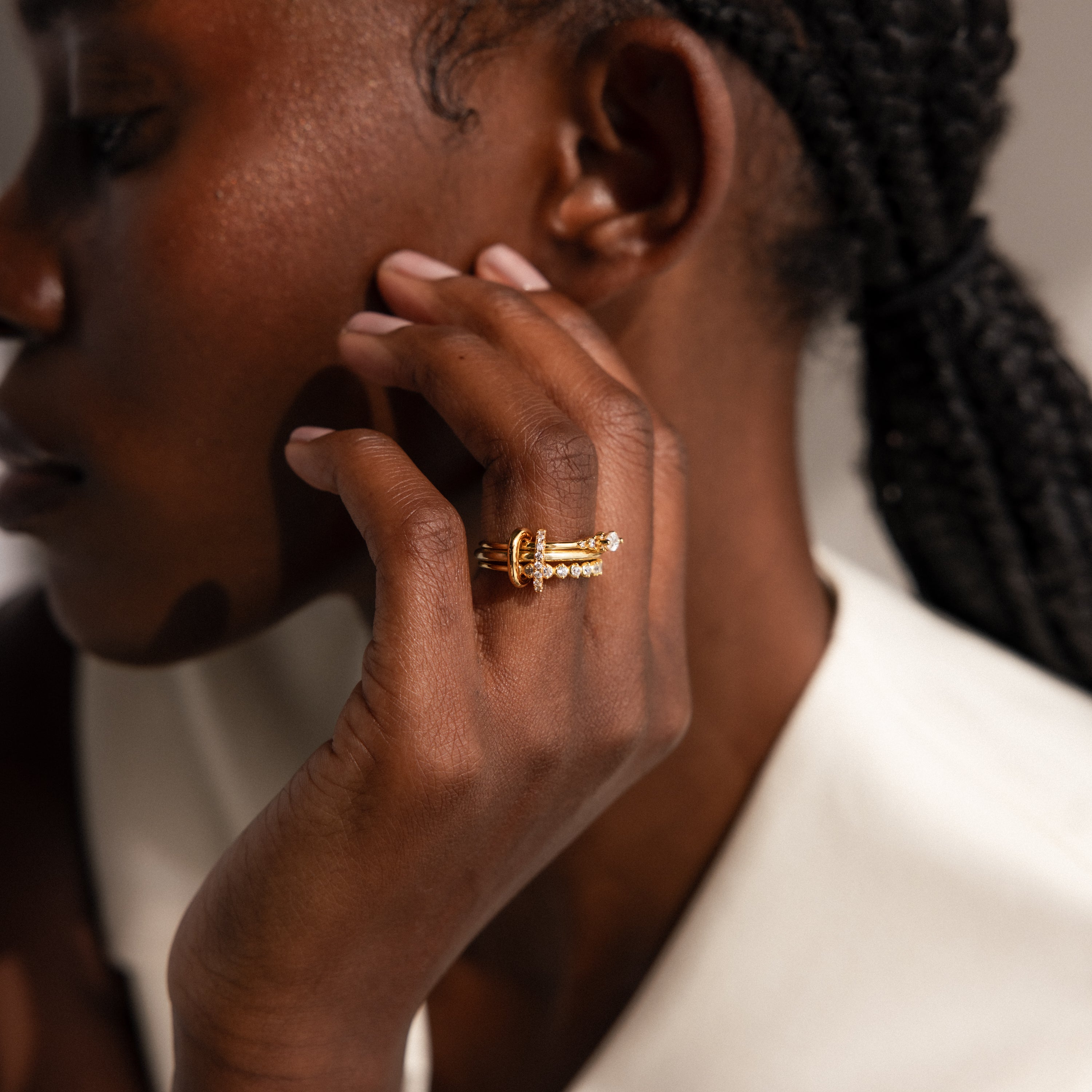 A person with braided hair wears gold rings, including the Multipurpose Pave Ring Lock Set featuring a pavé-detailed clasp, and rests their hand near their face in a white top—showcasing unique jewelry customization.