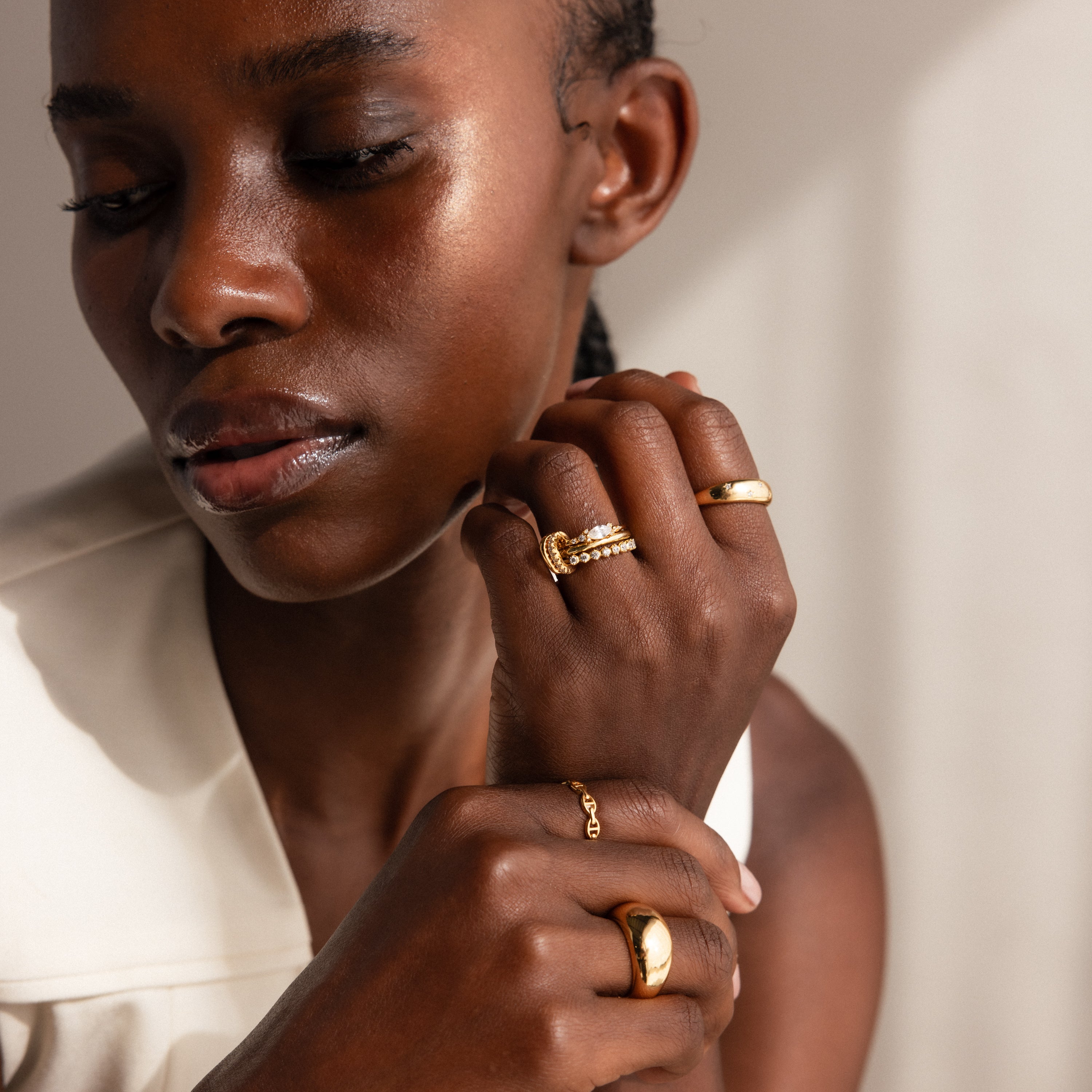A woman with closed eyes, wearing a cream-colored top, thoughtfully rests her chin on her hand adorned with multiple gold rings, including the Multipurpose Pave Ring Lock Set in 18K Gold featuring a pavé-detailed clasp.