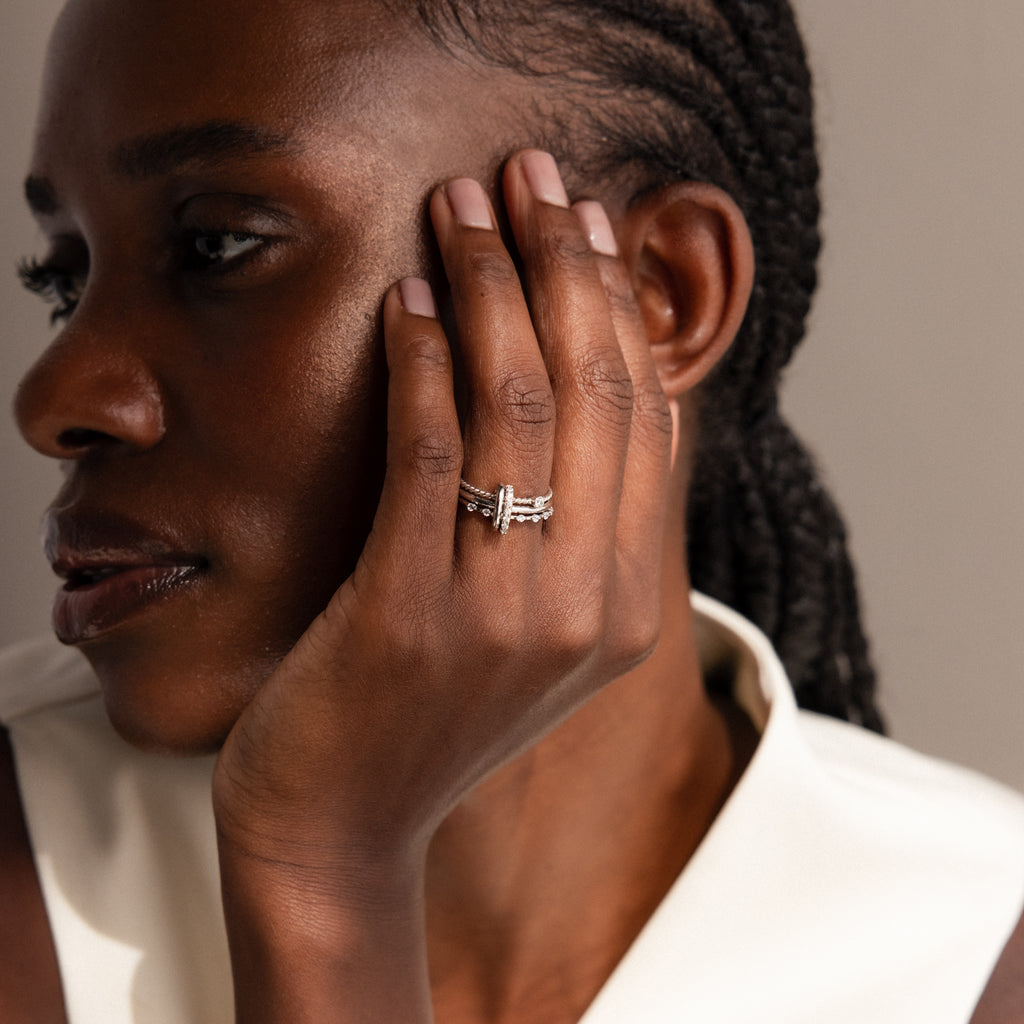 A woman with braids touches her face, showcasing stacked rings and the Multipurpose Pave Ring Lock Set in White Gold on her fingers, while wearing a white top.
