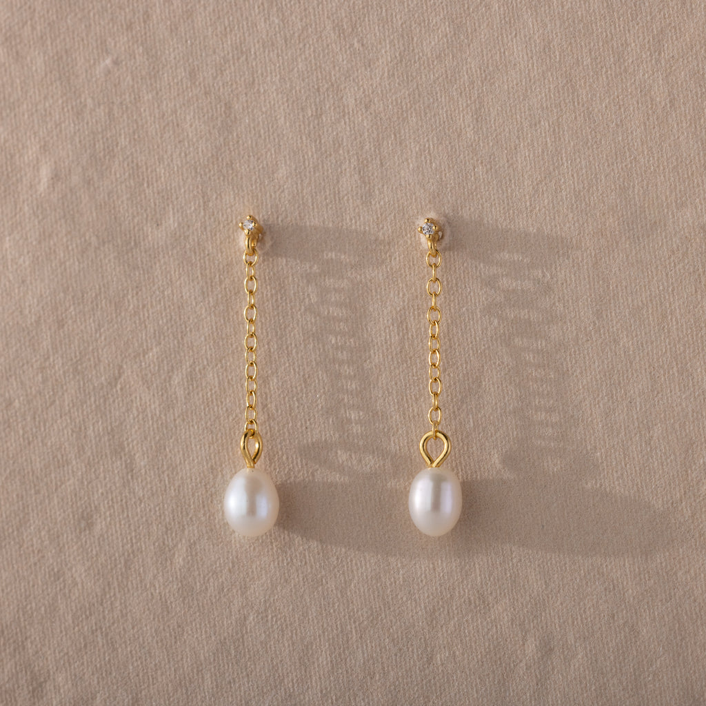 The Isha Pearl Drop Earrings showcase gold chain drop styling and elegant pearl charms, presented on a textured beige background.