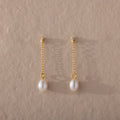 The Isha Pearl Drop Earrings showcase gold chain drop styling and elegant pearl charms, presented on a textured beige background.