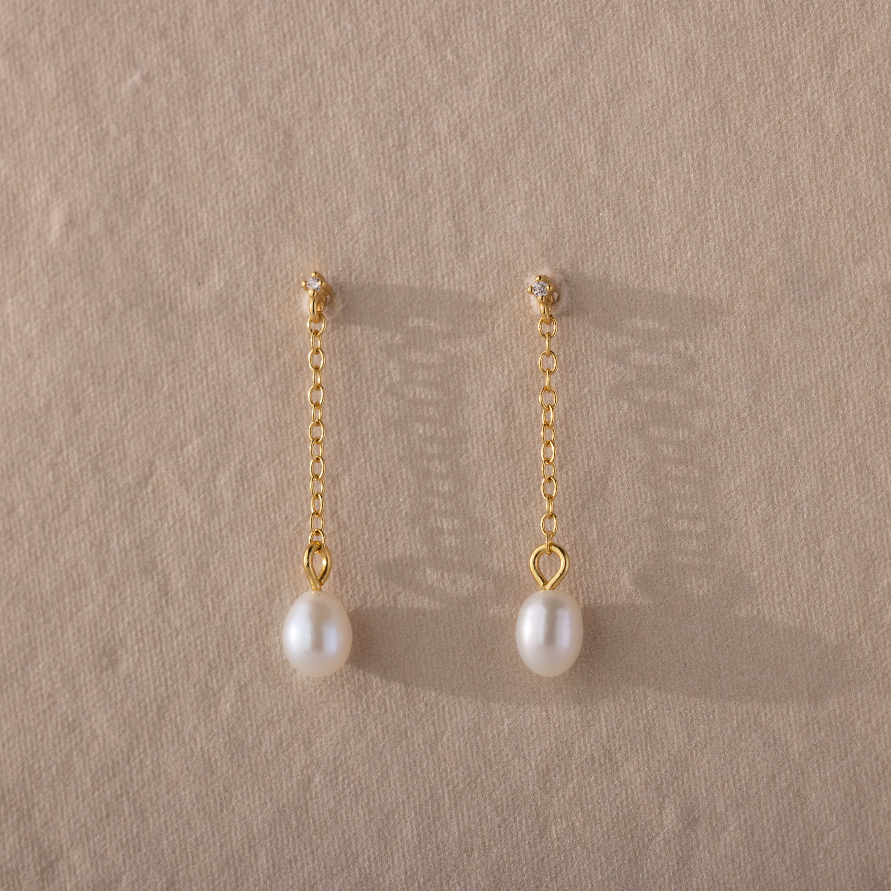 The Isha Pearl Drop Earrings showcase gold chain drop styling and elegant pearl charms, presented on a textured beige background.