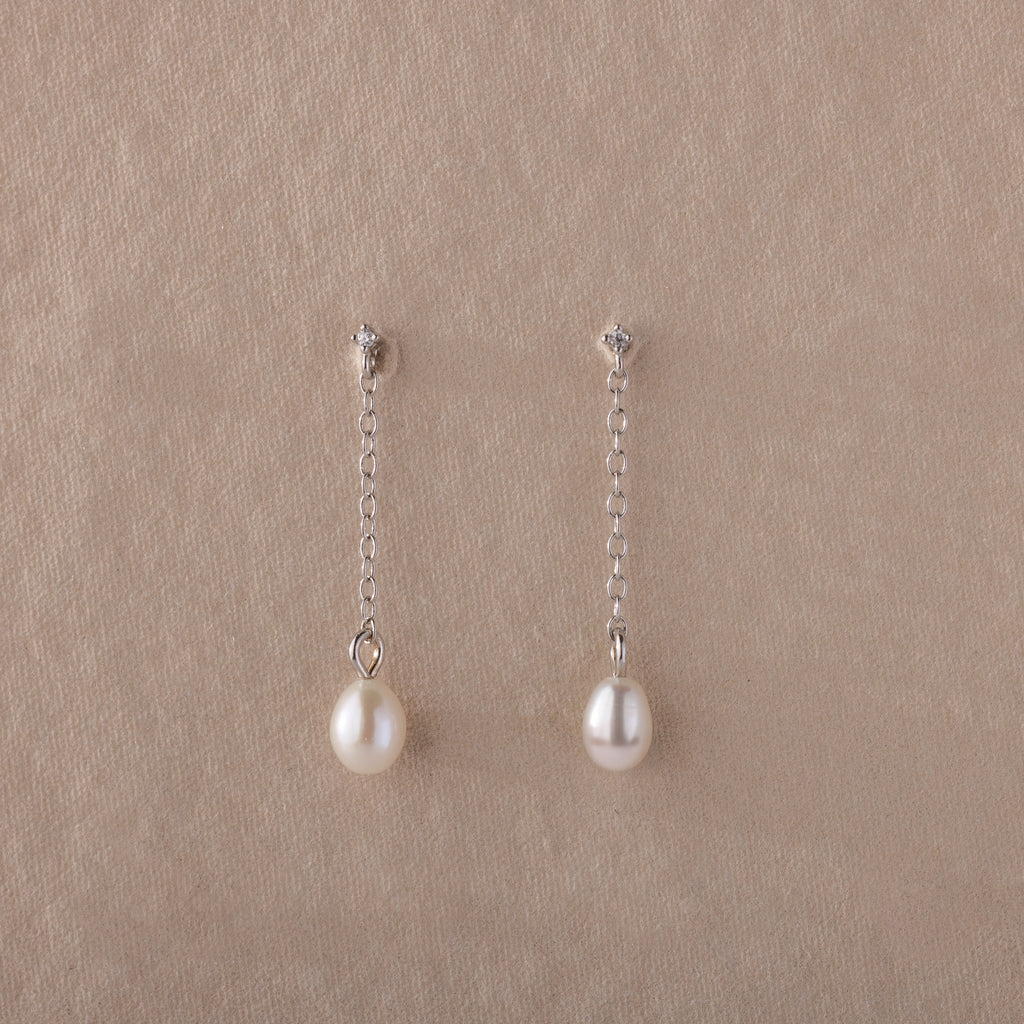 Isha Pearl Drop Earrings feature delicate pearls on silver chains, elegantly displayed against a beige textured background.