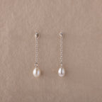 Isha Pearl Drop Earrings feature delicate pearls on silver chains, elegantly displayed against a beige textured background.