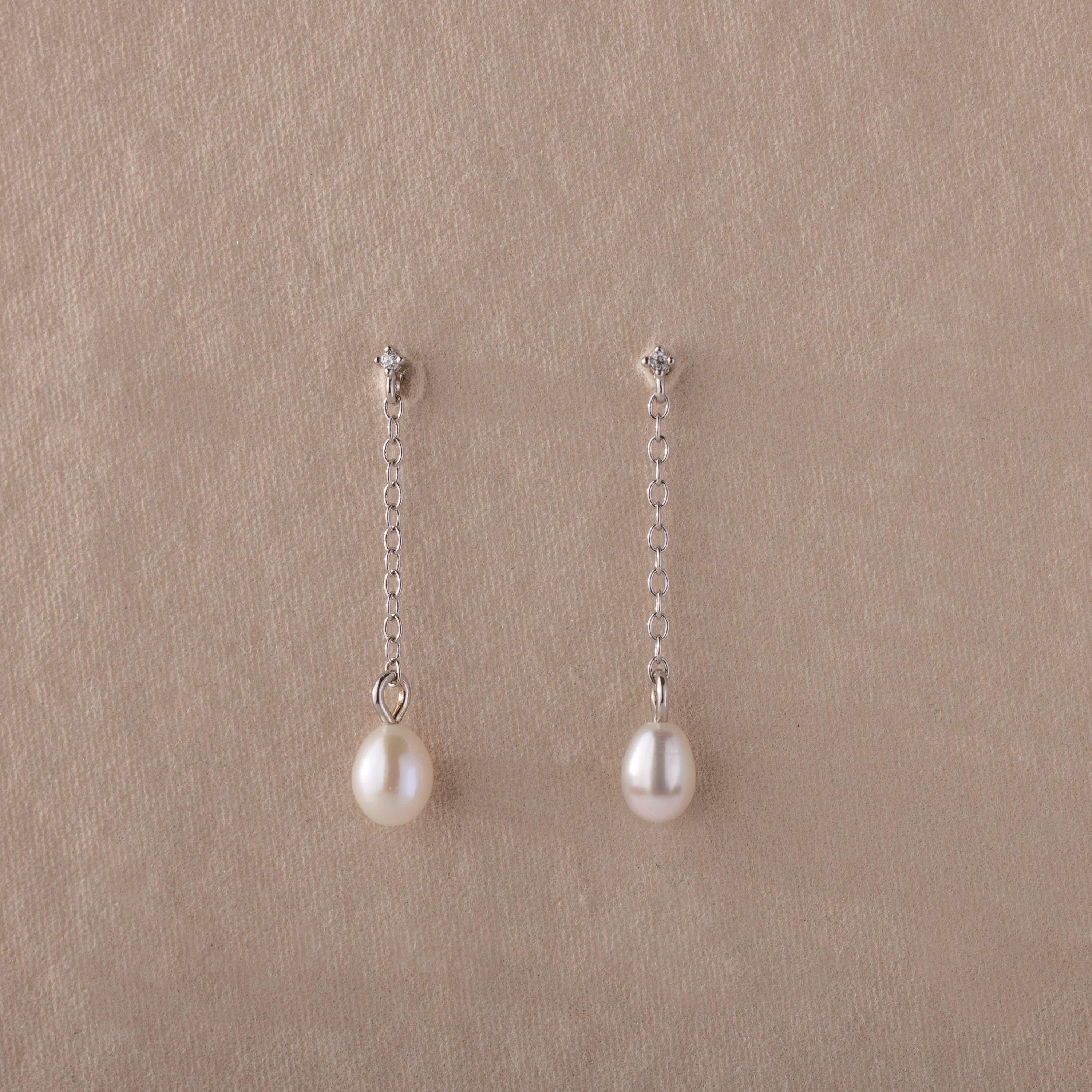 Isha Pearl Drop Earrings feature delicate pearls on silver chains, elegantly displayed against a beige textured background.