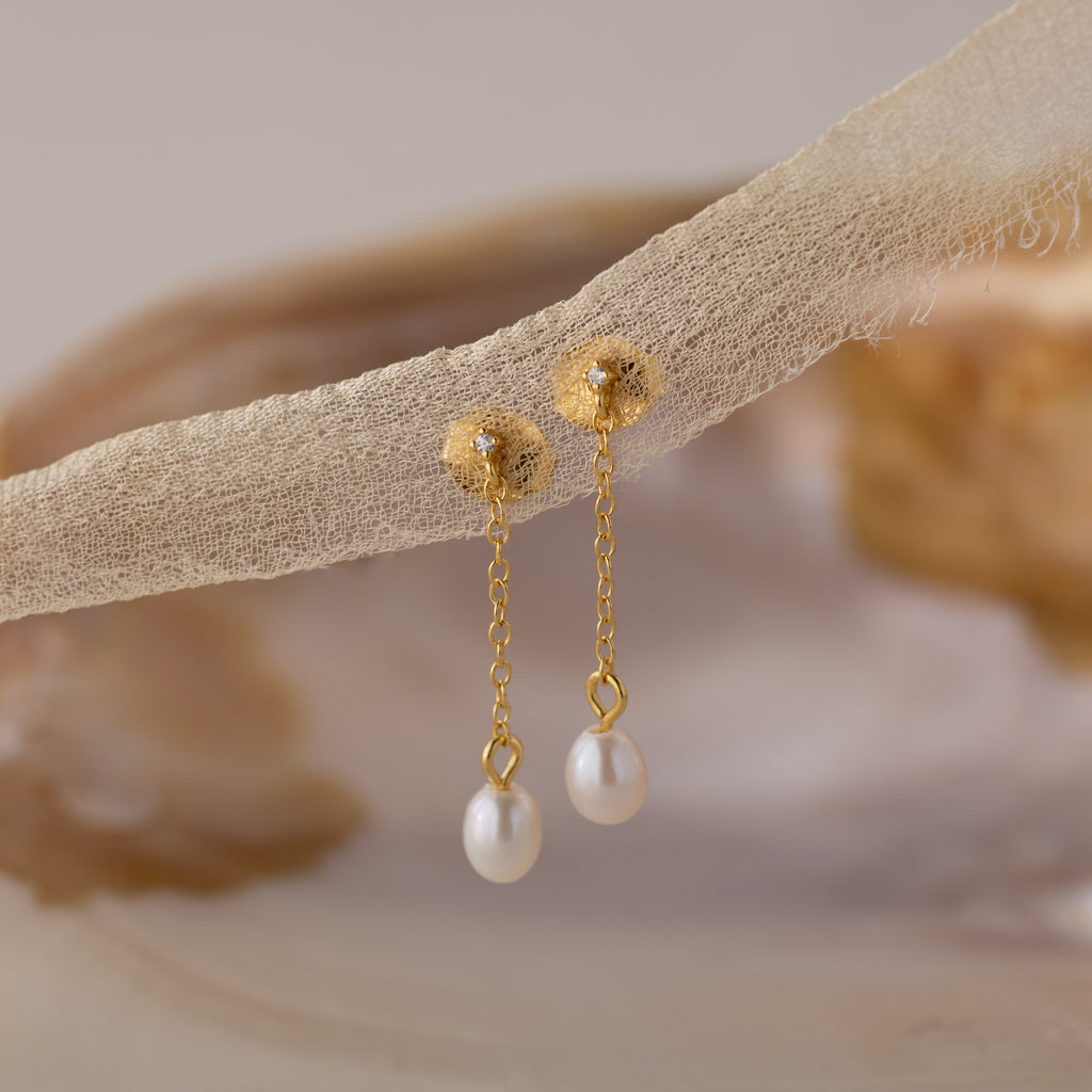 A pair of gold pearl drop earrings hanging from a sheer, textured fabric. Each earring features a textured gold stud with a small embedded crystal, a delicate chain, and a single white freshwater pearl dangling at the end