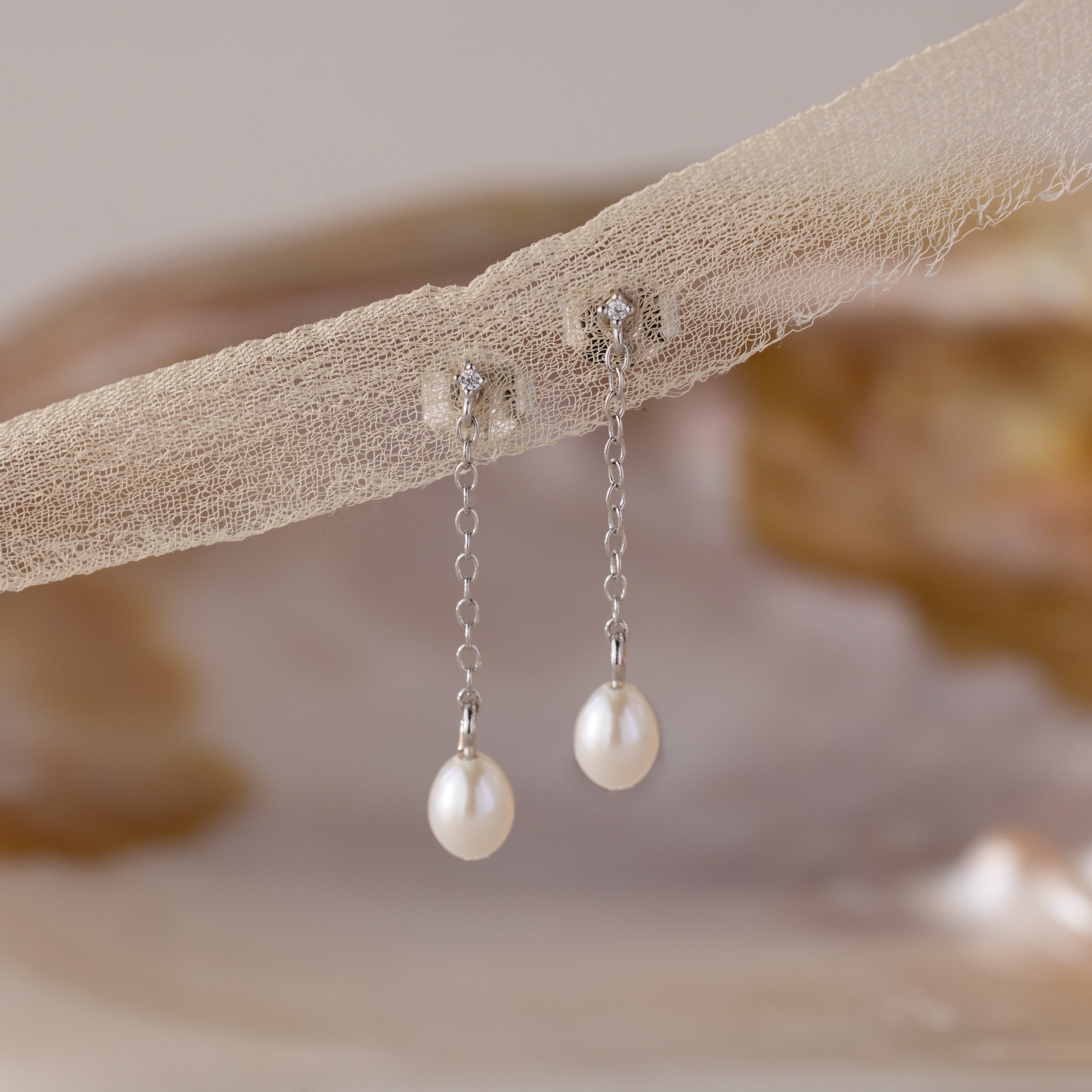 The Isha Pearl Drop Earrings showcase pearl charms and silver chains hanging gracefully from delicate beige fabric.
