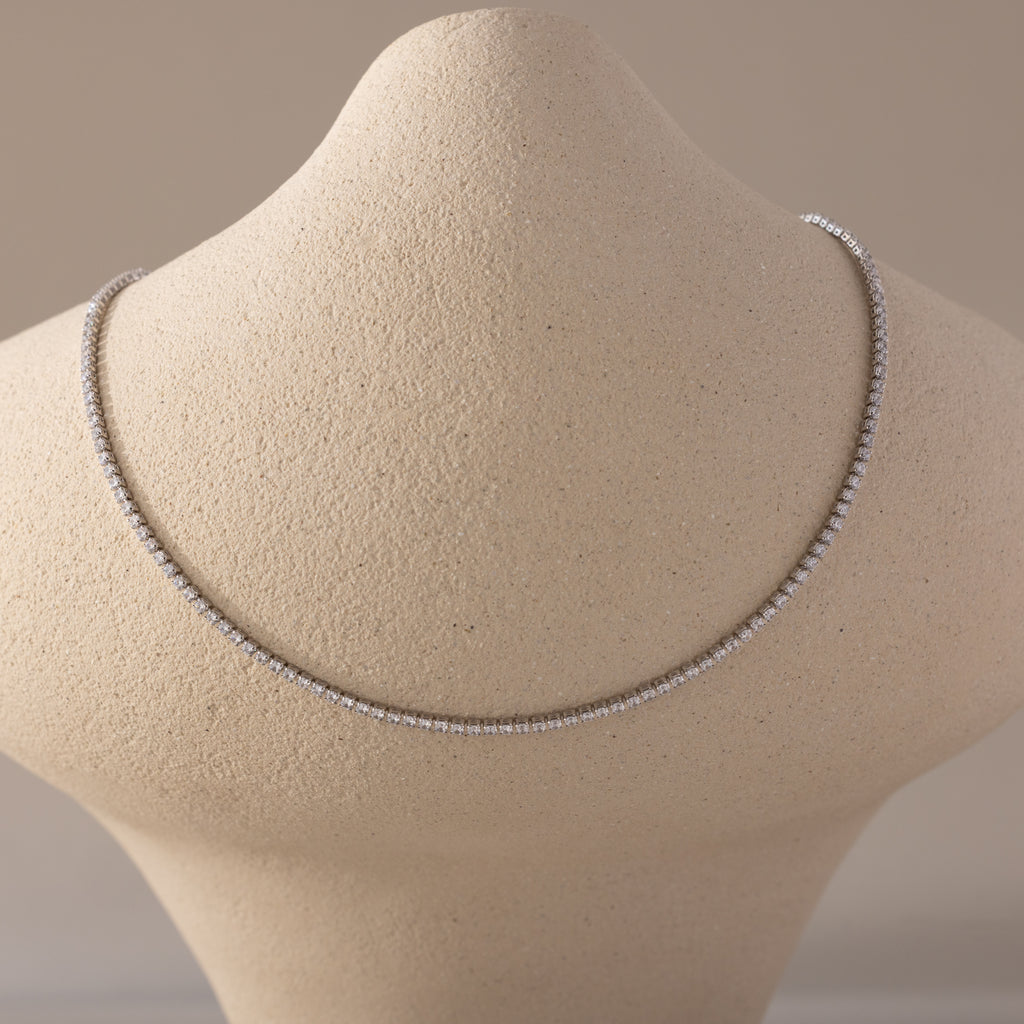  A Diamond Tennis Choker Necklace in white gold displayed on a beige textured neck form. The necklace features a continuous line of brilliant-cut white diamonds set in a sleek white gold setting, forming a minimalist and luxurious curve across the collarbone.