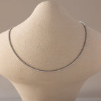  A Diamond Tennis Choker Necklace in white gold displayed on a beige textured neck form. The necklace features a continuous line of brilliant-cut white diamonds set in a sleek white gold setting, forming a minimalist and luxurious curve across the collarbone.
