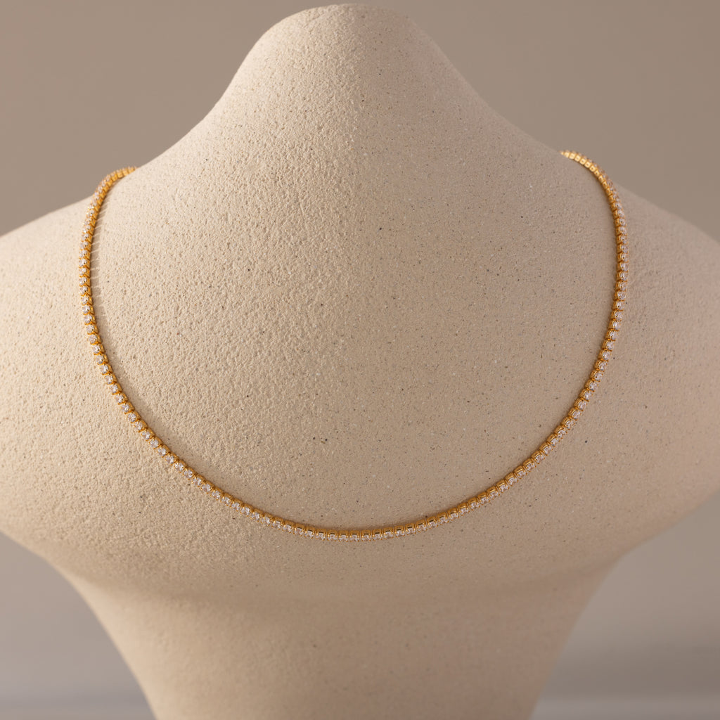  A close-up view of a Diamond Tennis Choker Necklace in 18K gold displayed on a textured beige neck form. The necklace features a continuous row of small, brilliant-cut diamonds set in gold, forming a delicate yet striking silhouette that rests elegantly along the collarbone line
