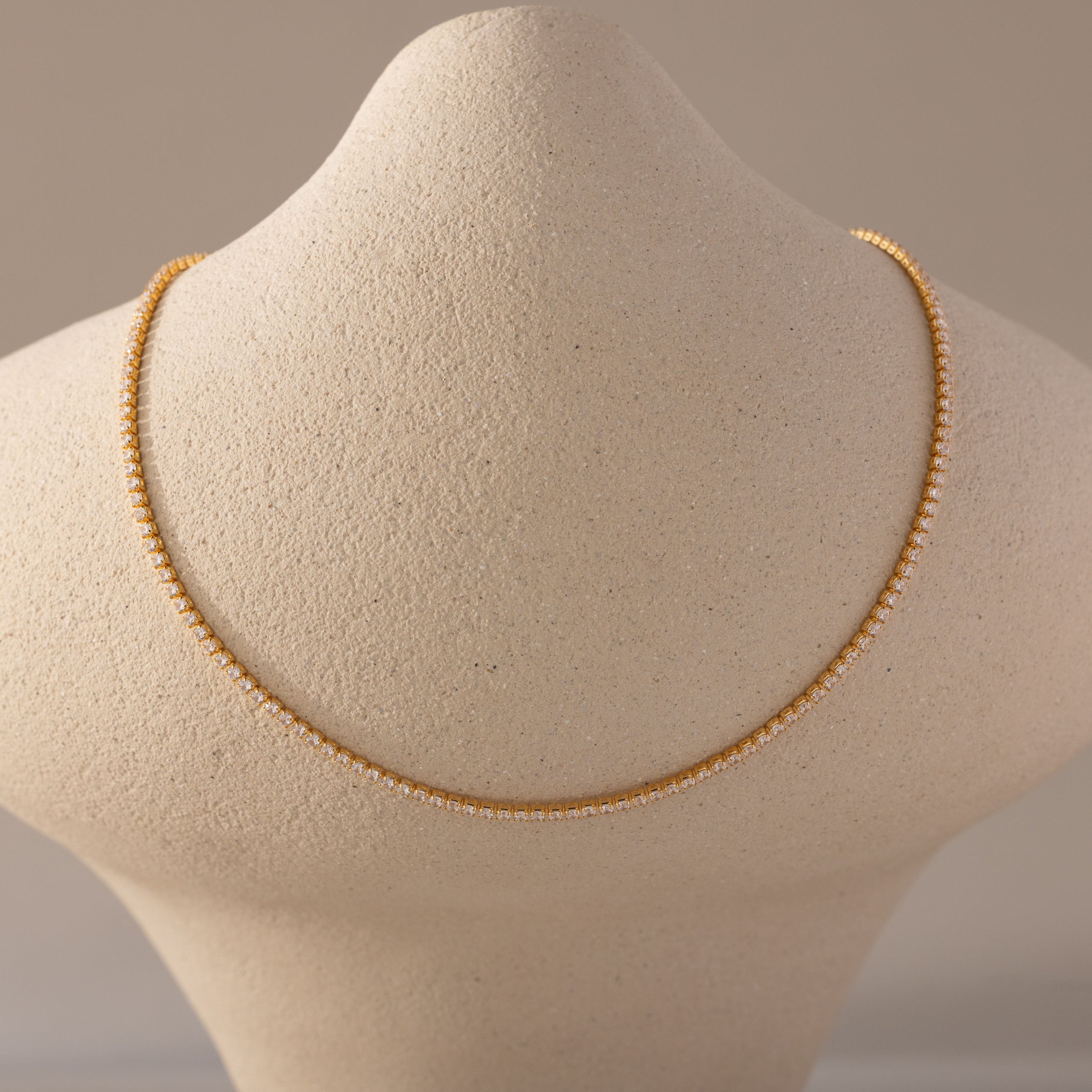  A close-up view of a Diamond Tennis Choker Necklace in 18K gold displayed on a textured beige neck form. The necklace features a continuous row of small, brilliant-cut diamonds set in gold, forming a delicate yet striking silhouette that rests elegantly along the collarbone line
