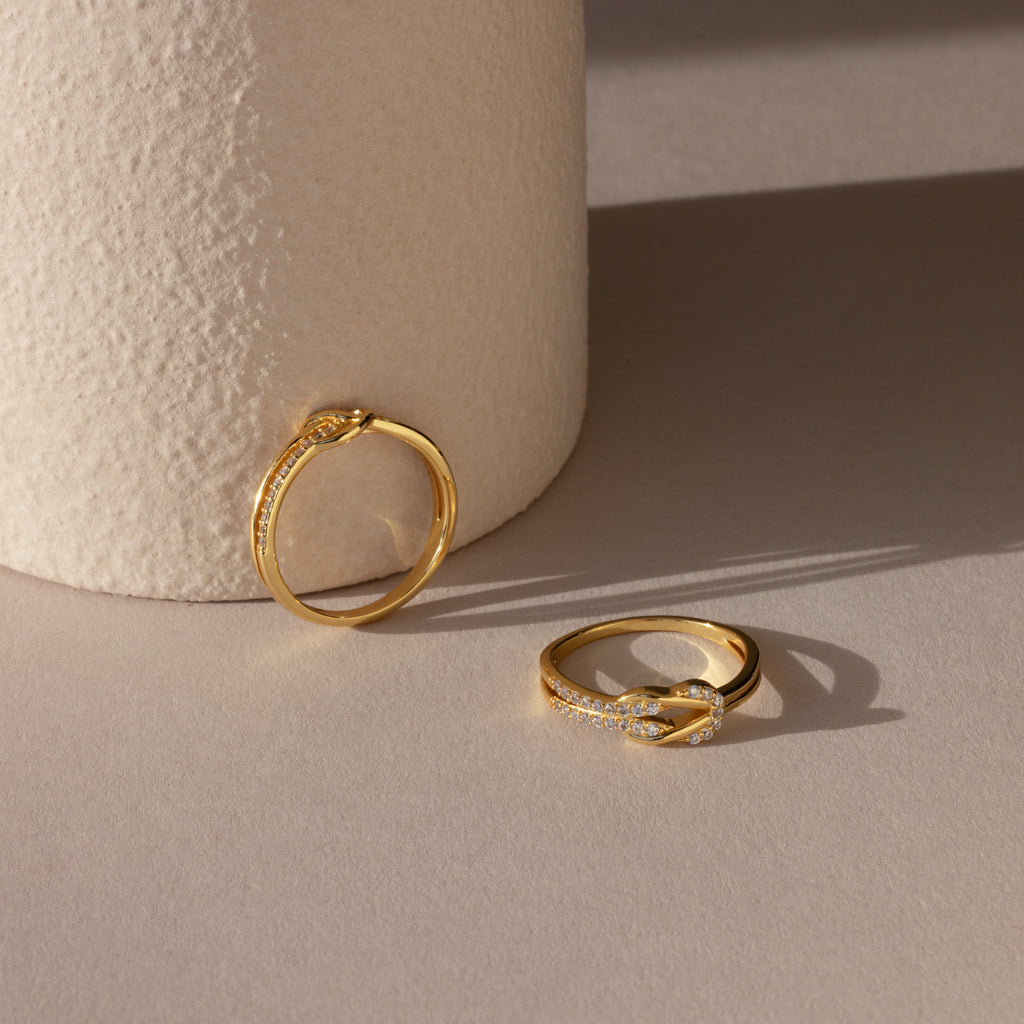 Two Pave Double Knot Rings with small diamonds rest on a light beige surface beside a textured cylinder—perfect examples of elegant knot jewelry.