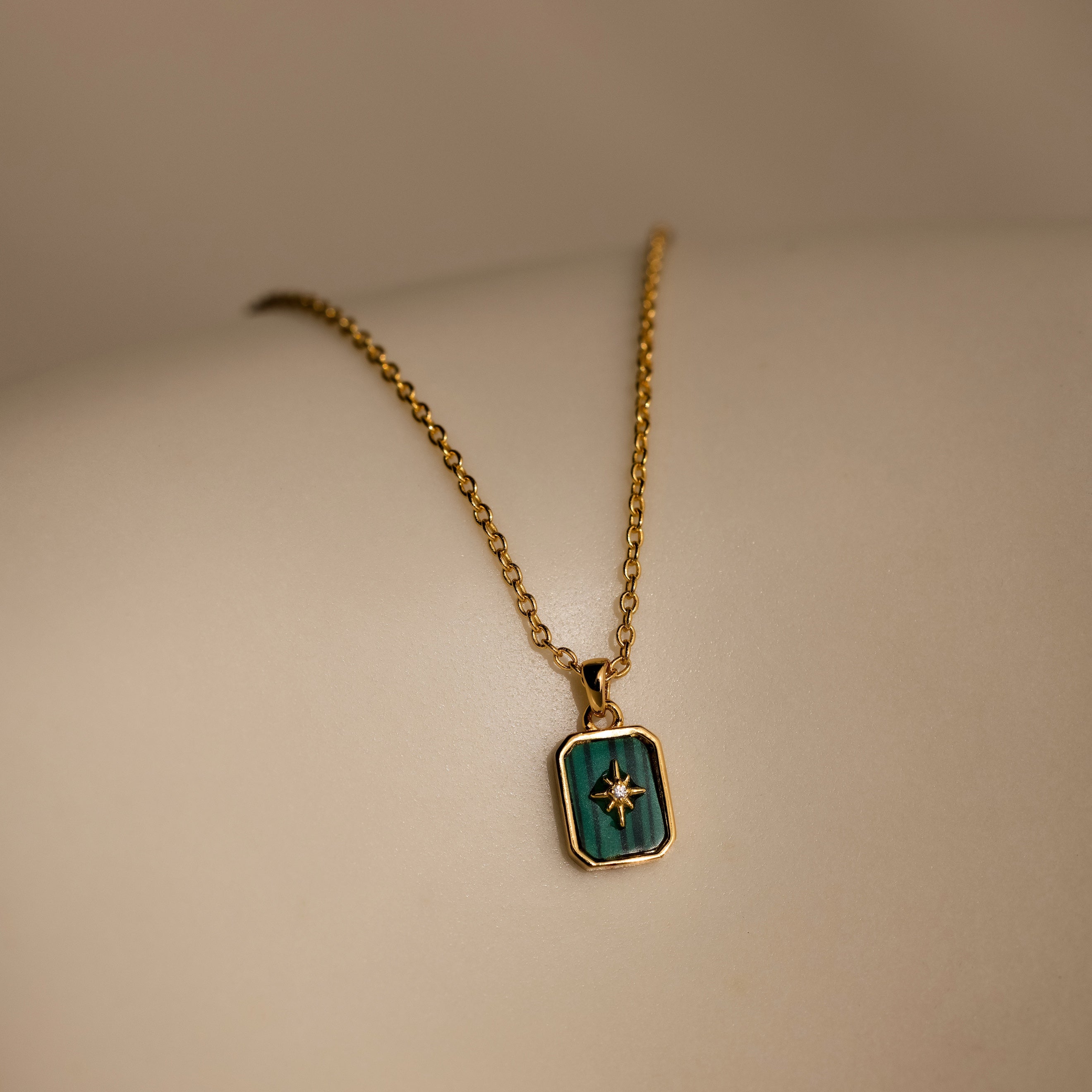 The North Star Gemstone Necklace in 18K Gold features a malachite rectangular pendant with a North Star design, displayed on a smooth surface.