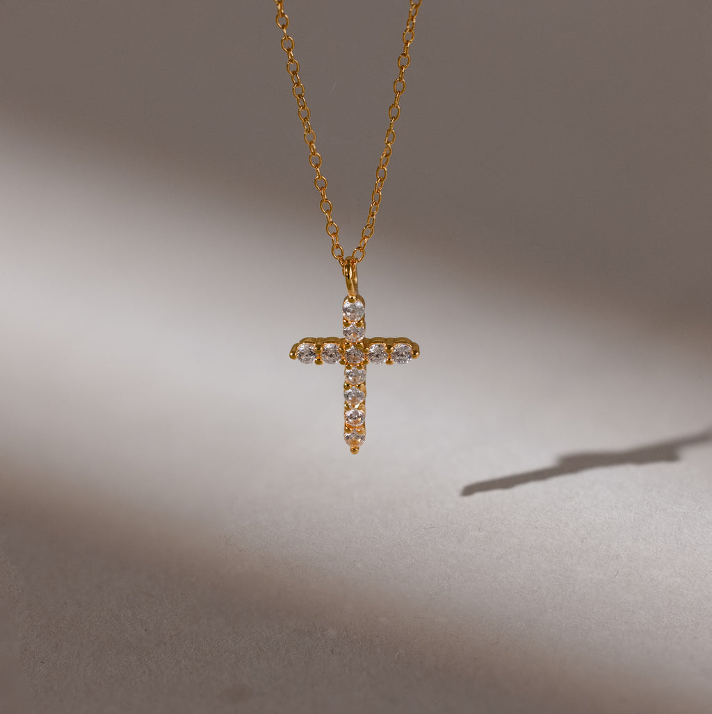 The Pave Cross Necklace in 18K Gold features a diamond-studded pendant casting a shadow on a neutral background—ideal for fans of elegant jewelry.