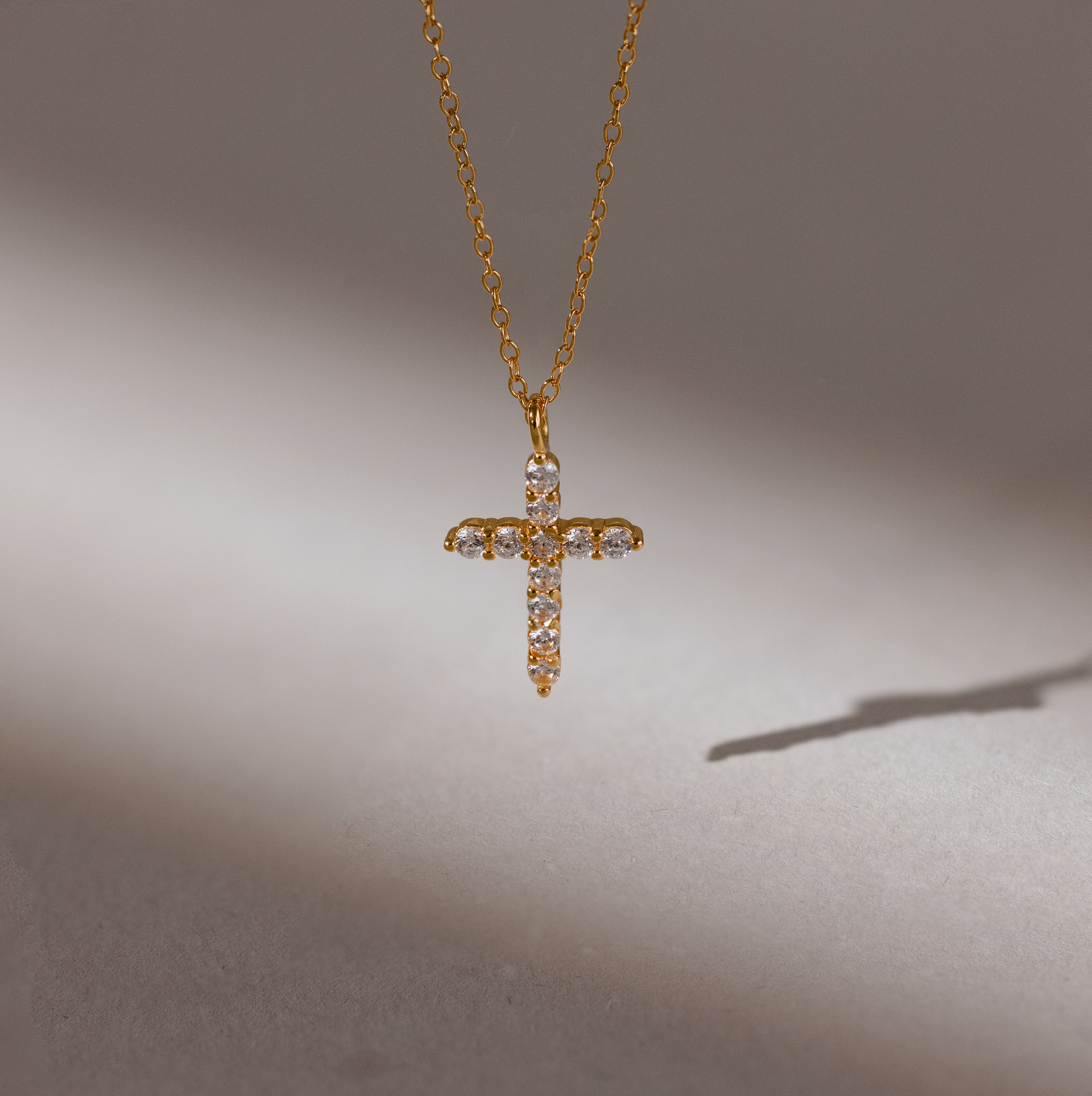 The Pave Cross Necklace in 18K Gold features a diamond-studded pendant casting a shadow on a neutral background—ideal for fans of elegant jewelry.