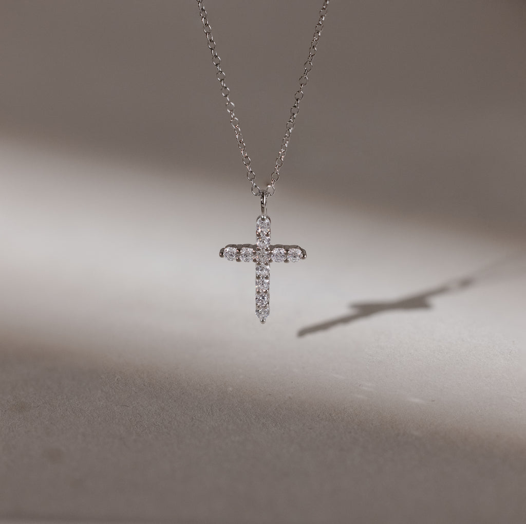 The Pave Cross Necklace in Silver features a sparkling pendant casting a shadow on a smooth surface—an exquisite piece of elegant jewelry.