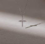 The Pave Cross Necklace shimmers with embedded diamonds, casting a delicate shadow on a smooth surface—an exquisite and elegant jewelry piece.