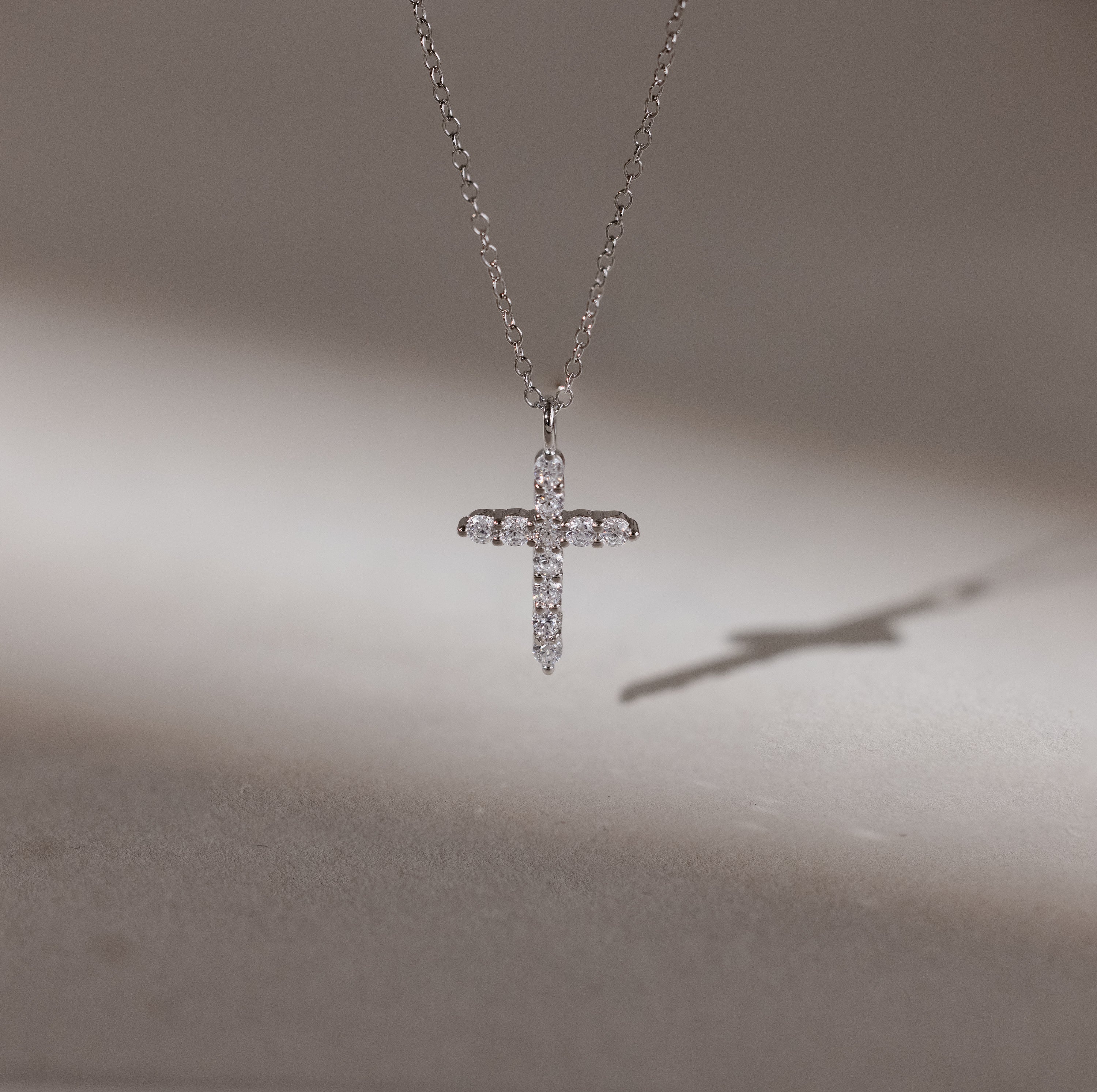The Pave Cross Necklace shimmers with embedded diamonds, casting a delicate shadow on a smooth surface—an exquisite and elegant jewelry piece.