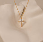 The Pave Cross Necklace features a gold chain and diamond-studded pendant, displayed on a cream surface—an elegant piece of jewelry perfect for any occasion.