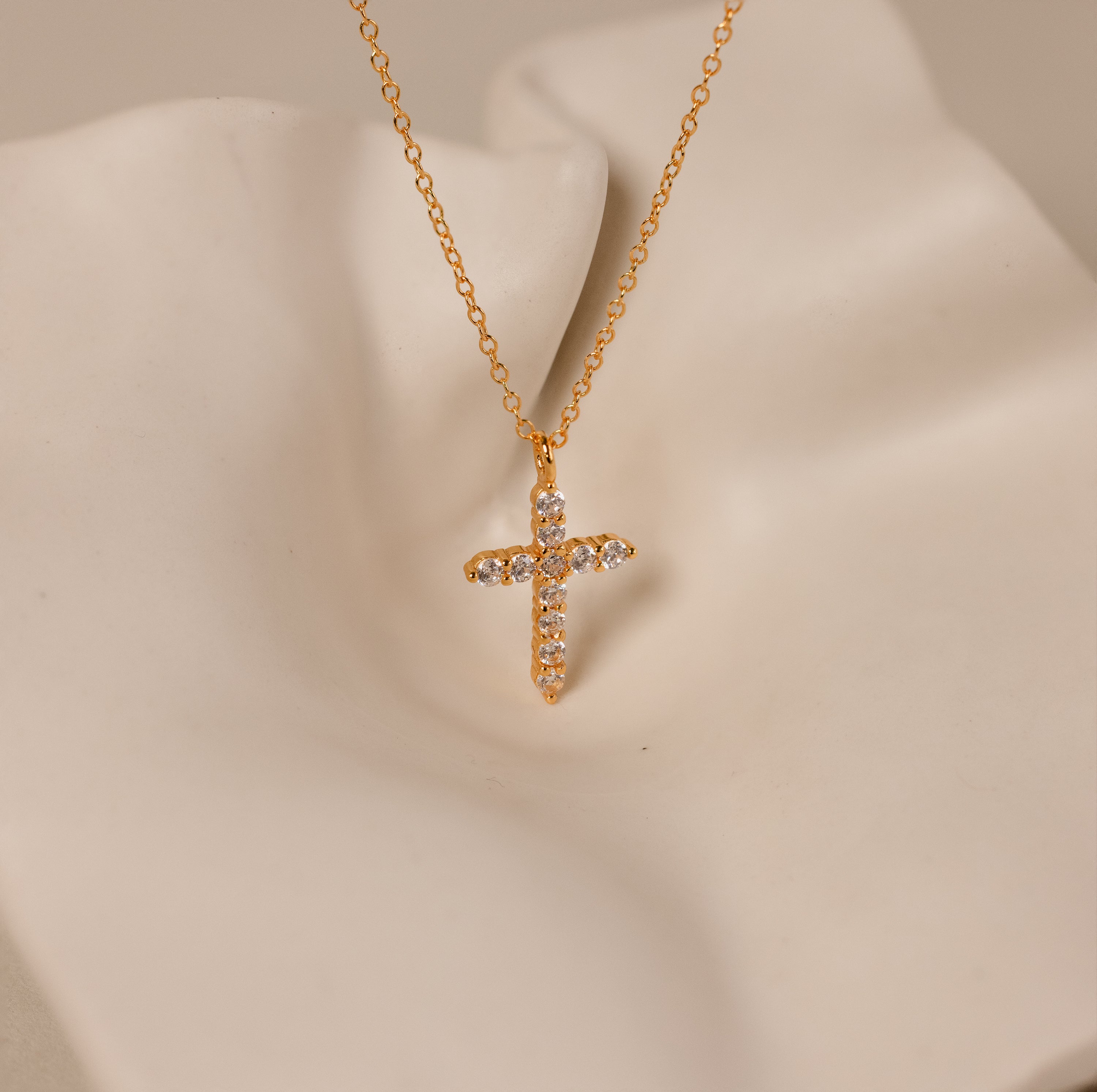 The Pave Cross Necklace features a gold chain and diamond-studded pendant, displayed on a cream surface—an elegant piece of jewelry perfect for any occasion.
