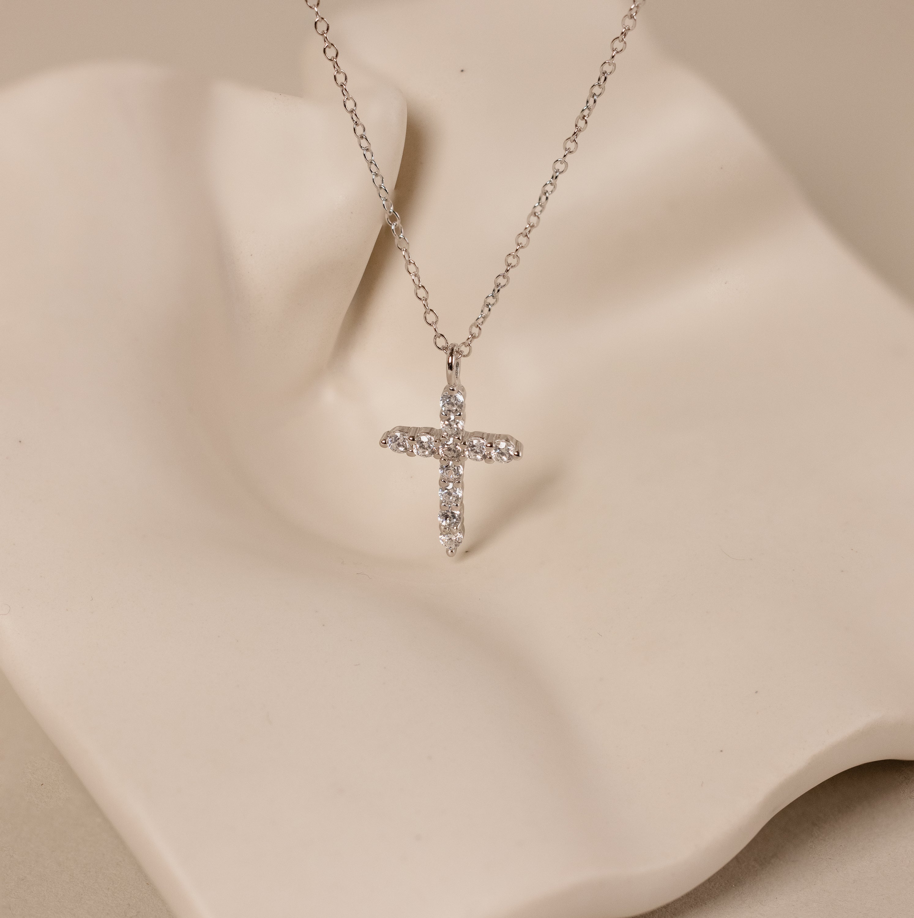 Discover elegance with the Pave Cross Necklace in Silver—a diamond-studded cross pendant on a sleek silver chain, beautifully showcased against a wavy cream-colored backdrop.