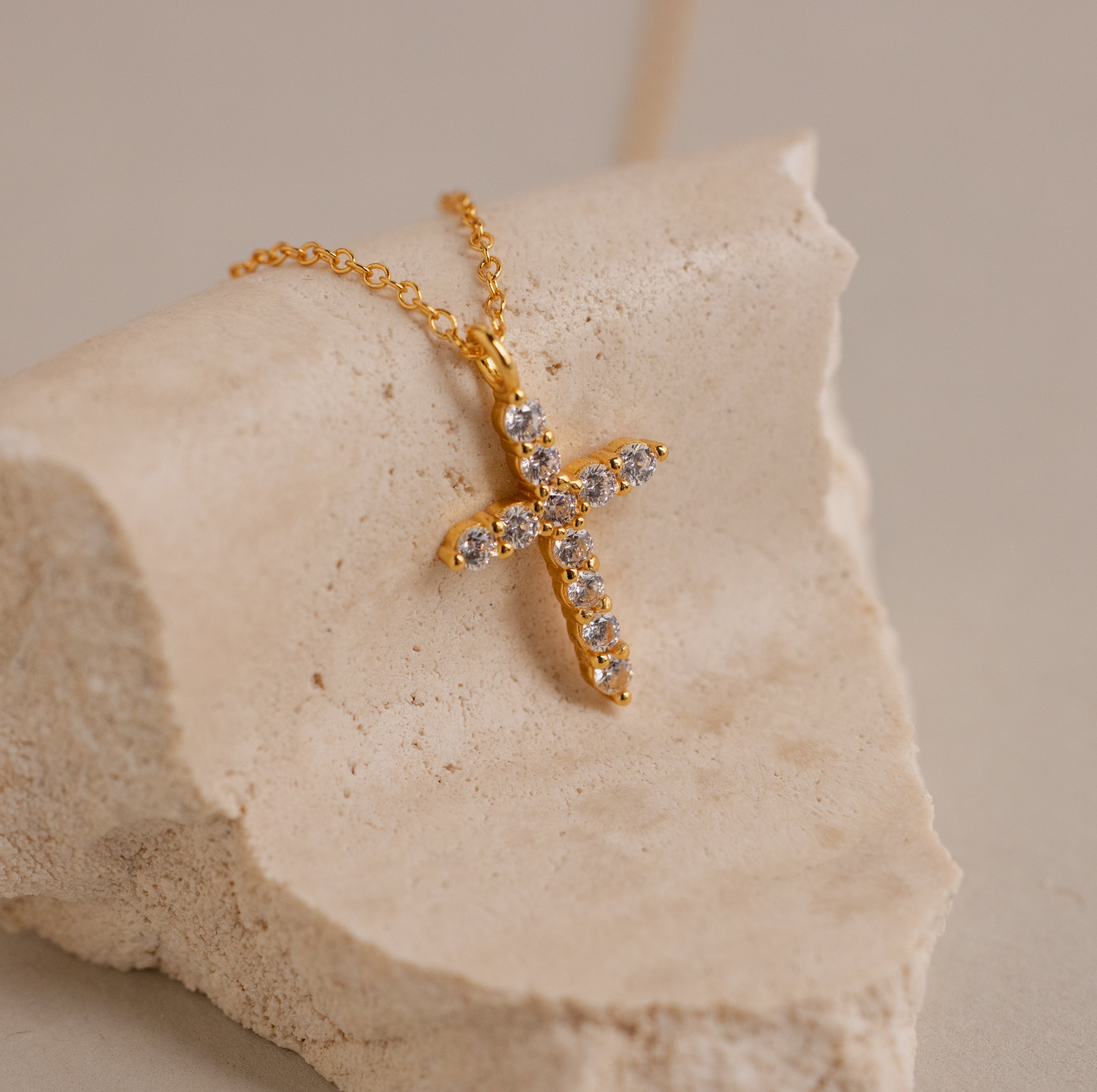 The Pave Cross Necklace features a gold cross pendant with shimmering gemstones on a gold chain, displayed on textured beige stone for an elegant touch.