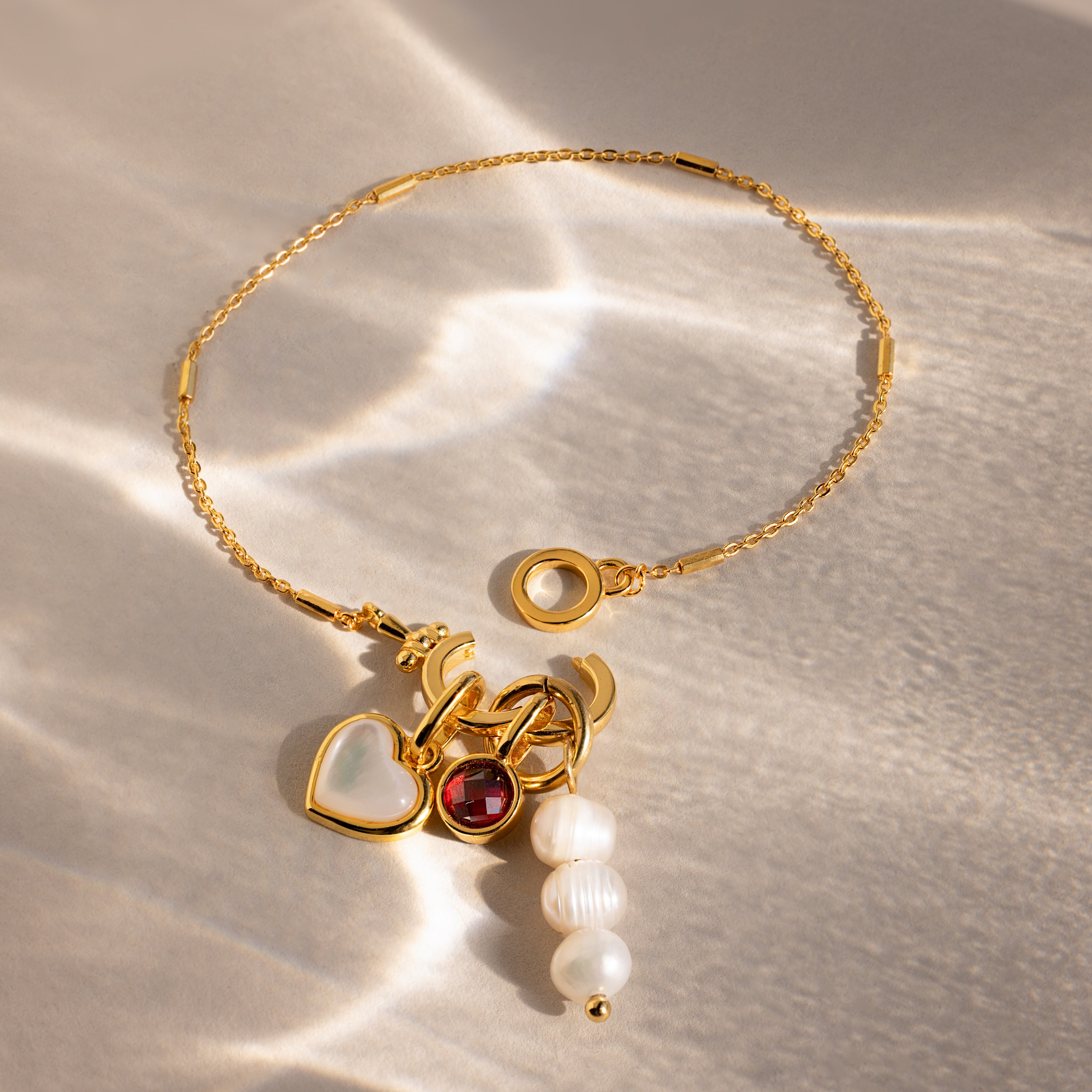 The Hinged Circle Charm Bracelet with heart, ruby, and pearl charms rests on a textured, sunlit surface—minimalist design meets elegant details.