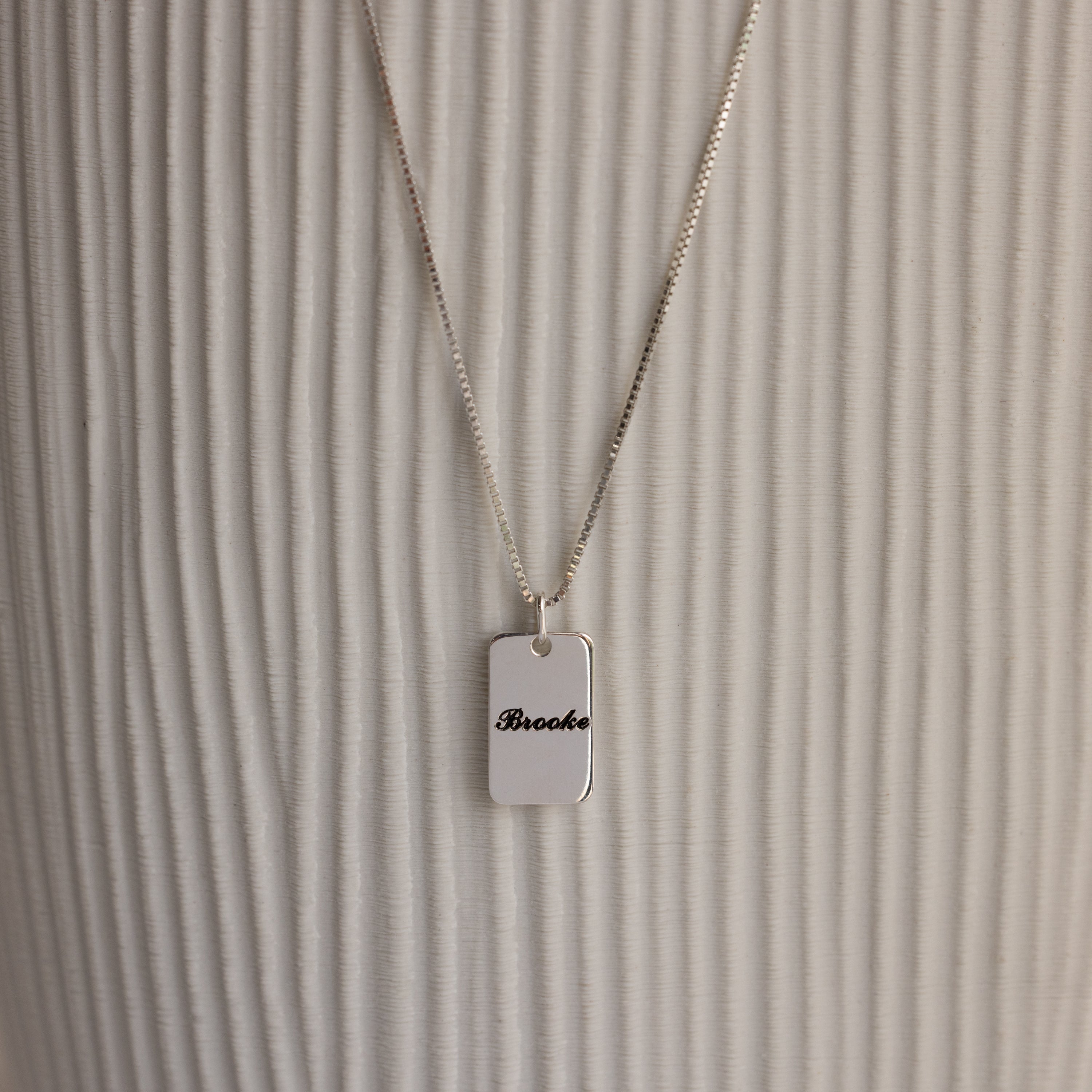 Name Tag Necklace in Sterling Silver with a rectangular pendant engraved "Brooke," shown on a textured white background.
