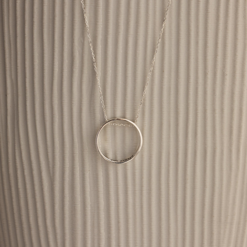 Inner Circle Necklace in Sterling Silver, featuring a simple circular pendant on a chain, shown against a textured beige background.